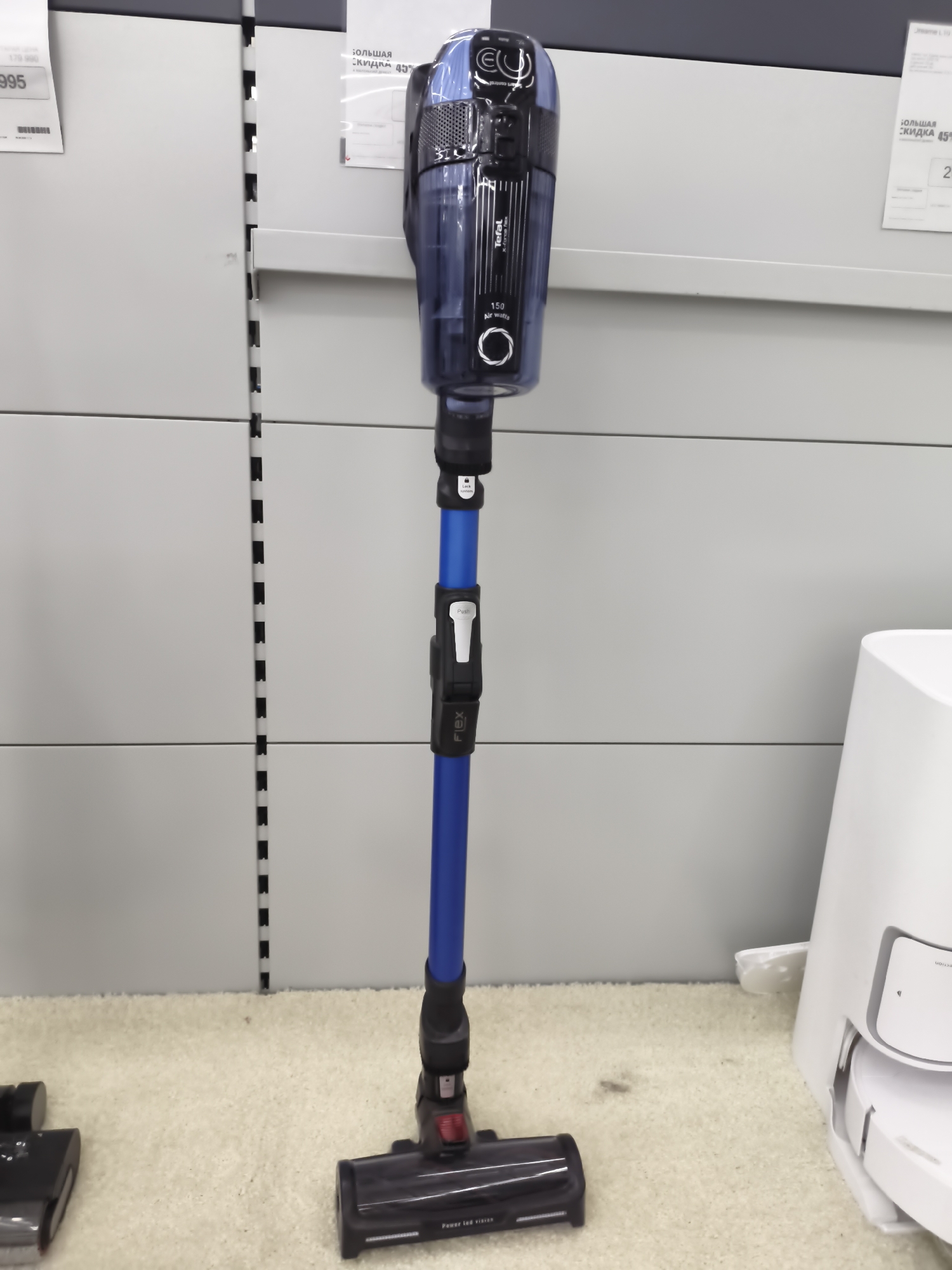 In Aktobe, Kazakhstan, an upright, blue and black cordless vacuum cleaner stands vertically on a beige carpeted floor. The vacuum has a transparent dustbin and a telescopic wand with a flex feature. At the base of the vacuum is the cleaning head. To the left of the main vacuum, a portion of another cleaning head is visible, suggesting a second vacuum cleaner is present, though its main body is not in frame. In the background, white shelving units are visible, with price tags attached to some of the shelves. One price tag displays the number "995", and another indicates a "45% discount." To the right of the vacuum, a portion of a white object, possibly a charging station or another appliance, is visible. The lighting in the scene is bright and even, typical of an indoor retail environment. There are no people visible in the image.