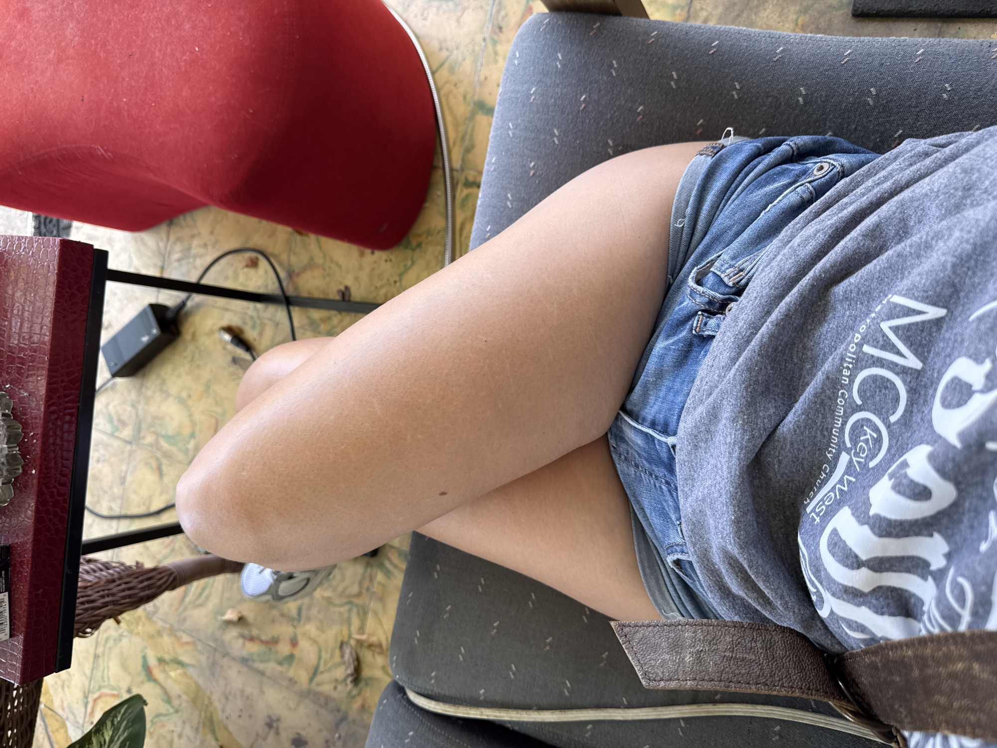 The image shows a close-up view of a person's legs and torso, seated in a chair. The person is wearing denim shorts and a gray t-shirt with white text. The t-shirt appears to be from a "Key West Metropolitan Community Church". The scene is indoors, with a red object and a wicker basket visible in the background. The floor has a patterned design. The lighting suggests it could be daytime. The perspective is from above, looking down at the person. The person's legs are crossed, and the focus is on their clothing and skin.