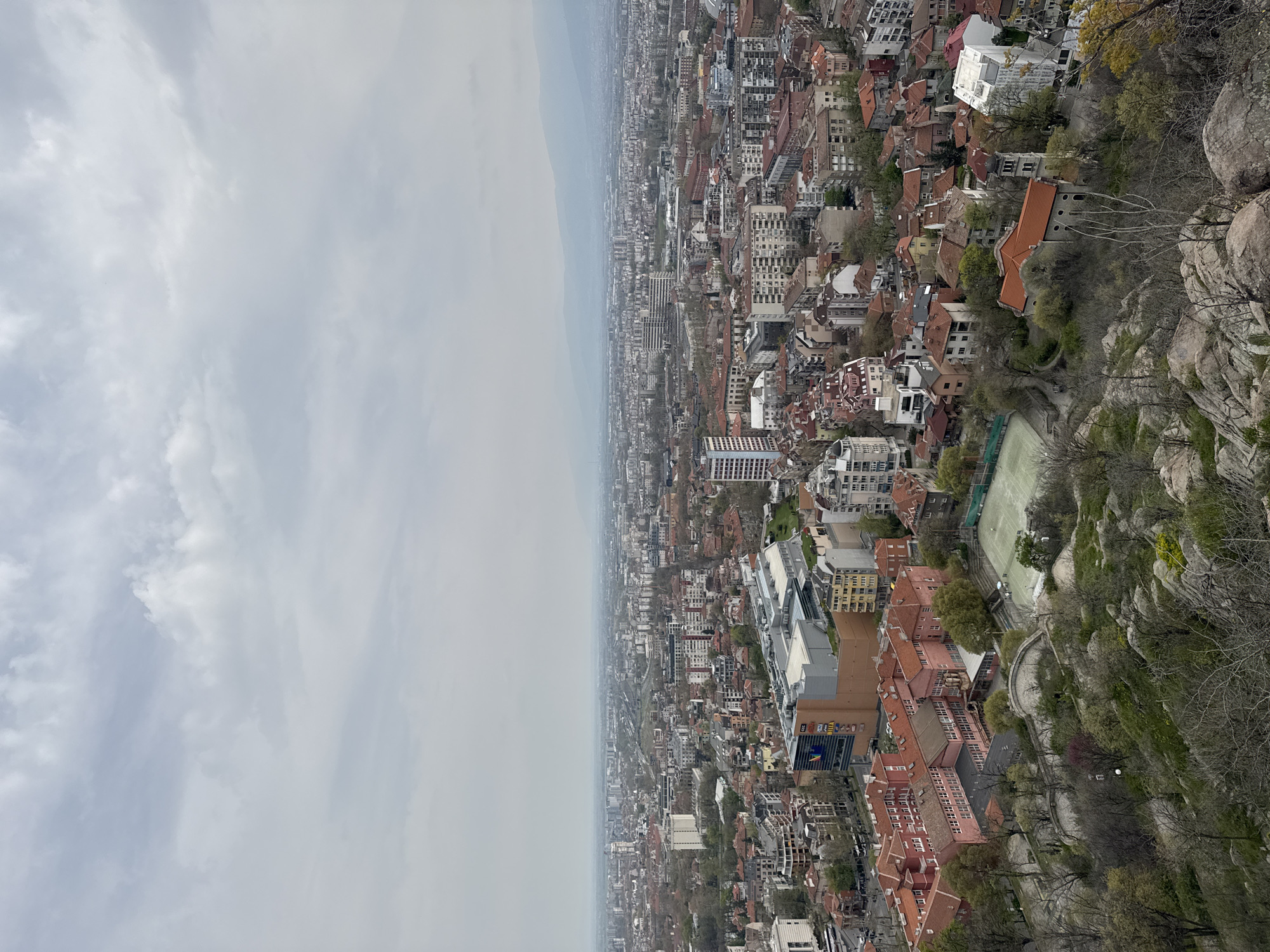 This is an aerial view of the city of Plovdiv, Bulgaria, taken from the Nebet Tepe hill. The image showcases the city's landscape, including the urban area with its buildings, structures, and infrastructure. The sky is overcast. Towards the right of the frame are rocky formations and sparse vegetation, transitioning into residential buildings with red-tiled roofs. A green-surfaced sports field is visible. Beyond the immediate buildings, the city extends into the distance with densely packed structures fading into a hazy horizon.