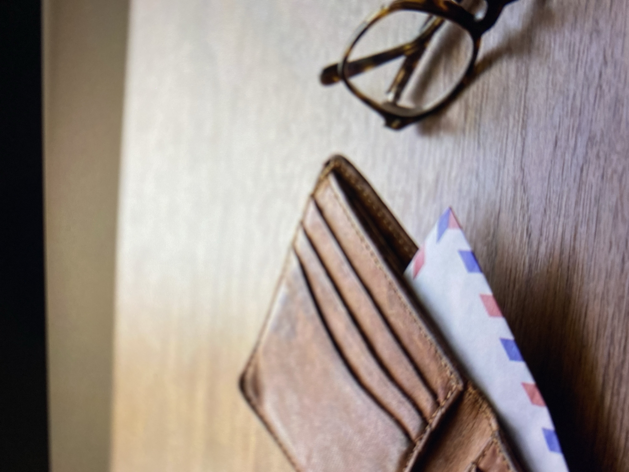A close-up, eye-level shot shows a brown leather wallet with multiple card slots, slightly ajar, on a wooden surface. An airmail envelope with a red, white, and blue diagonal stripe pattern peeks out from beneath the wallet. To the right of the wallet, a pair of tortoiseshell glasses with rounded frames rests on the wooden surface. The background is a blurred out dark surface. The image does not depict the city of Kuta Selatan, Indonesia.