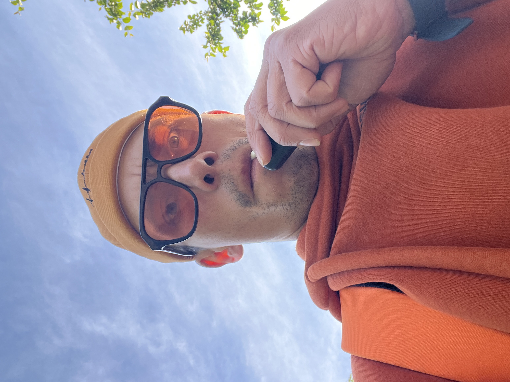 An upward-angled shot captures an individual looking down towards the camera, set against a bright, clear blue sky with sparse, wispy clouds. The person, appearing to be an adult male, is prominently featured in the foreground, taking up a significant portion of the frame.
He is wearing a light brown or tan beanie/hood and sunglasses with distinctly orange-tinted lenses, which reflect some light, making the eyes partially visible behind them. There is visible stubble on his face. His right hand is raised, holding a small, dark, cylindrical object, possibly a vape pen or similar electronic device, positioned close to his mouth. He is dressed in an orange or reddish-brown hooded sweatshirt or hoodie, with the fabric folds visible on his upper body. A dark-colored watch or wristband is visible on his right wrist.
In the upper background, branches with green leaves from a tree or bush are visible, suggesting an outdoor setting. The bright lighting indicates it is daytime, likely a sunny day with good weather. There are no clearly legible texts visible on the clothing or accessories, though a faint, dark pattern or logo is present on the beanie/hood. The overall impression is a casual outdoor moment. This scene takes place in Chaikovsky, Russia.