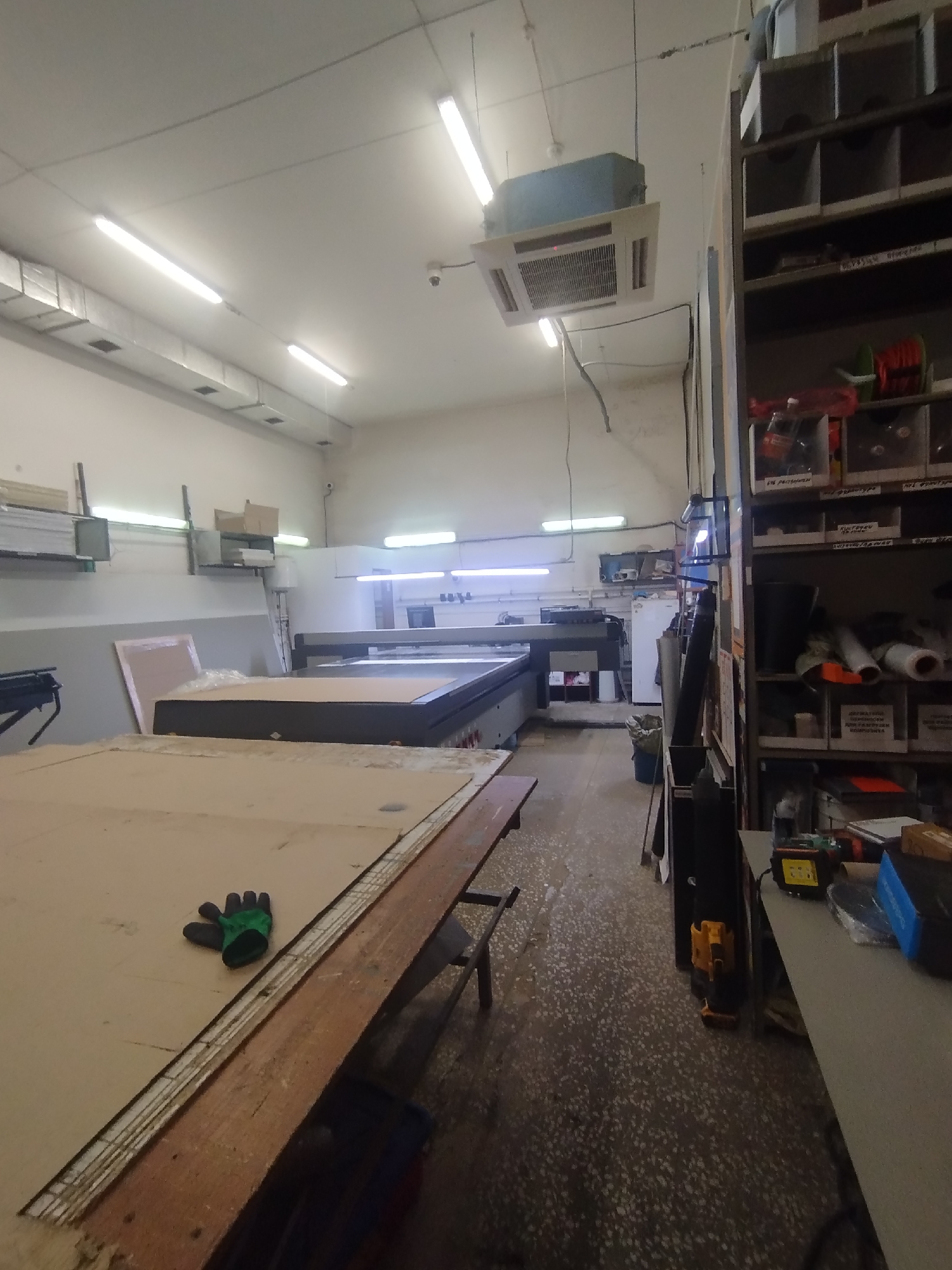 The image depicts an indoor industrial workshop or storage area, likely located in Naberezhnye Chelny, Russia. The scene is filled with various equipment and materials associated with manufacturing or fabrication. Dominating the foreground is a large, flat surface covered with cardboard, with a green work glove lying on it. To the left, a portion of a workbench or table with a wooden edge is visible. In the mid-ground, a large, flatbed machine, possibly a CNC router or a large format printer, is situated, with a pinkish panel resting against it. Further back, industrial lighting fixtures are mounted on the ceiling, along with air conditioning units and ductwork. To the right, a tall shelving unit is filled with various items, including rolls of material, containers, and boxes, suggesting organized storage. The floor is a speckled, grey concrete surface, showing signs of wear. The overall impression is of an active workspace, possibly involved in the production of signage, large-format printing, or similar industrial processes.