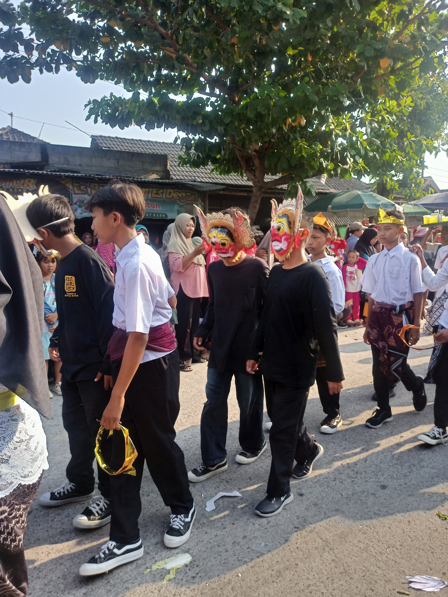 A group of young males wearing decorative masks are walking in a procession along a paved street in Saren, Indonesia. The masks appear to be elaborately decorated with bright colors and stylized features, resembling animal or mythical creatures. Some of the individuals are also wearing traditional attire, such as sarongs and gold crowns. They are walking on a street lined with buildings and trees, suggesting a public event or festival is taking place. The sunlight indicates it is daytime, casting shadows on the ground. The background includes a building with a sign that reads "ES Degan" and a tree with lush green foliage. Other people, including women and children, can be seen observing the procession in the background. The overall scene depicts a cultural or religious gathering, likely a parade or celebration.