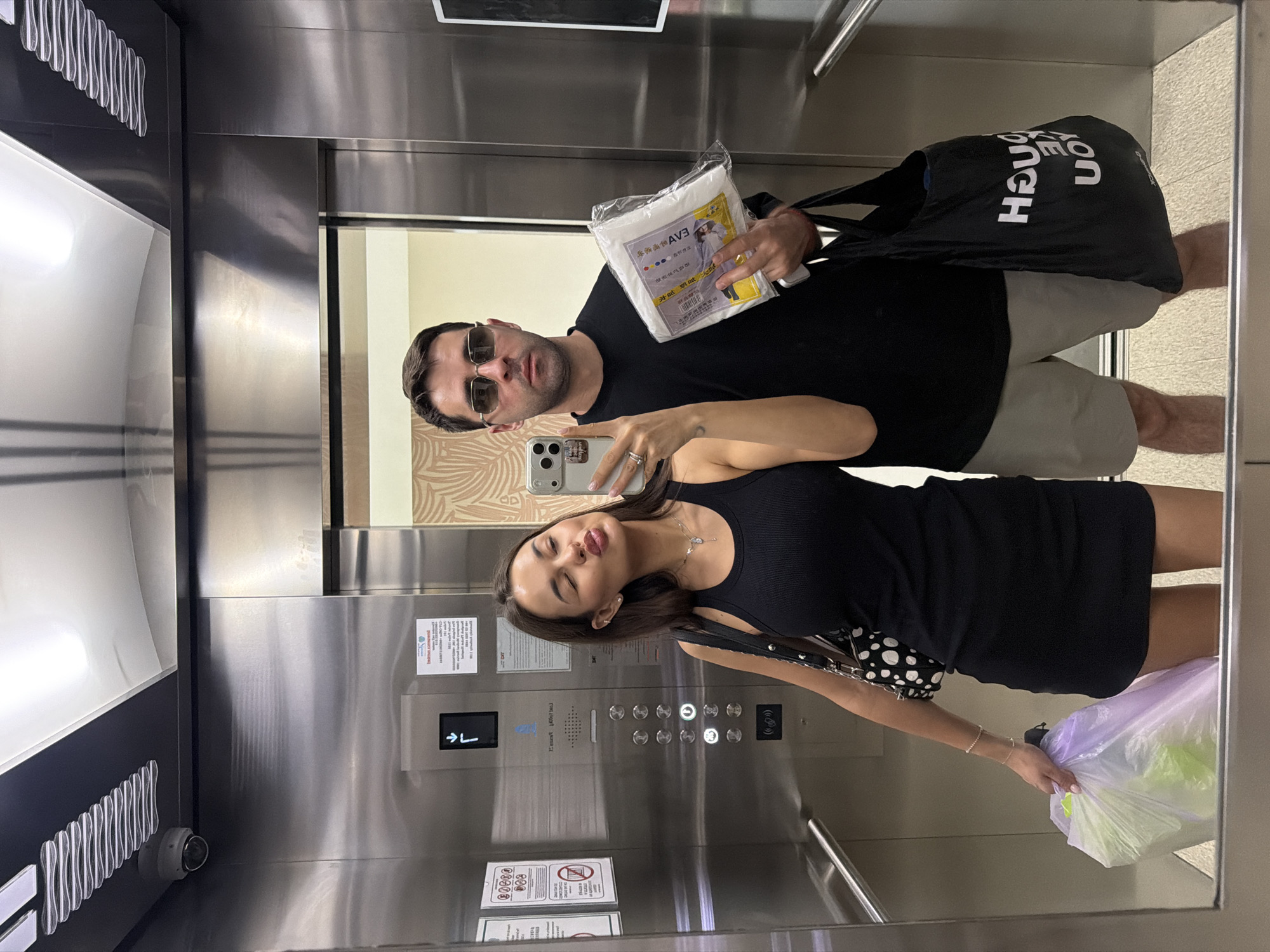 The photo depicts a couple in an elevator. The man is wearing sunglasses and a black shirt and is holding a package of items with Chinese text. The woman is wearing a black tank top and skirt, holding a shopping bag. The elevator has a silver control panel with buttons and information labels, along with safety notices. The surroundings suggest an indoor setting, possibly in Choeng Thale, Thailand, considering the presence of Thai and Chinese language. A security camera is visible in the elevator.