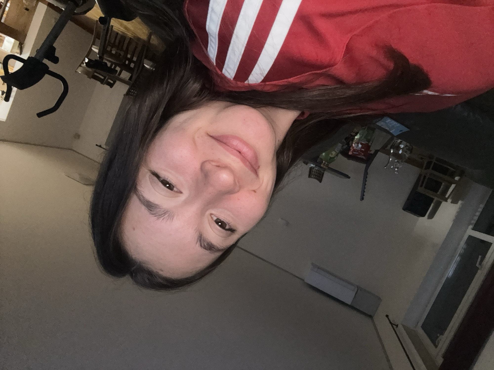 A young woman with dark hair and brown eyes smiles at the camera. She is wearing a red shirt with white stripes. The background is an indoor setting with a window, a table, and exercise equipment. The location is specified as the city of Sofia, Bulgaria.