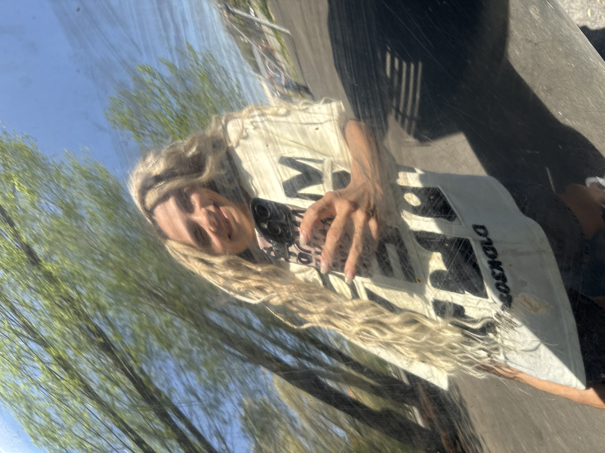 The image shows a woman taking a selfie in a reflective surface, likely a car hood. The reflection shows trees with green leaves and a clear blue sky, suggesting an outdoor location. The woman has long blonde hair and is wearing a white t-shirt with black text. The location is Rusanovo, Russia.