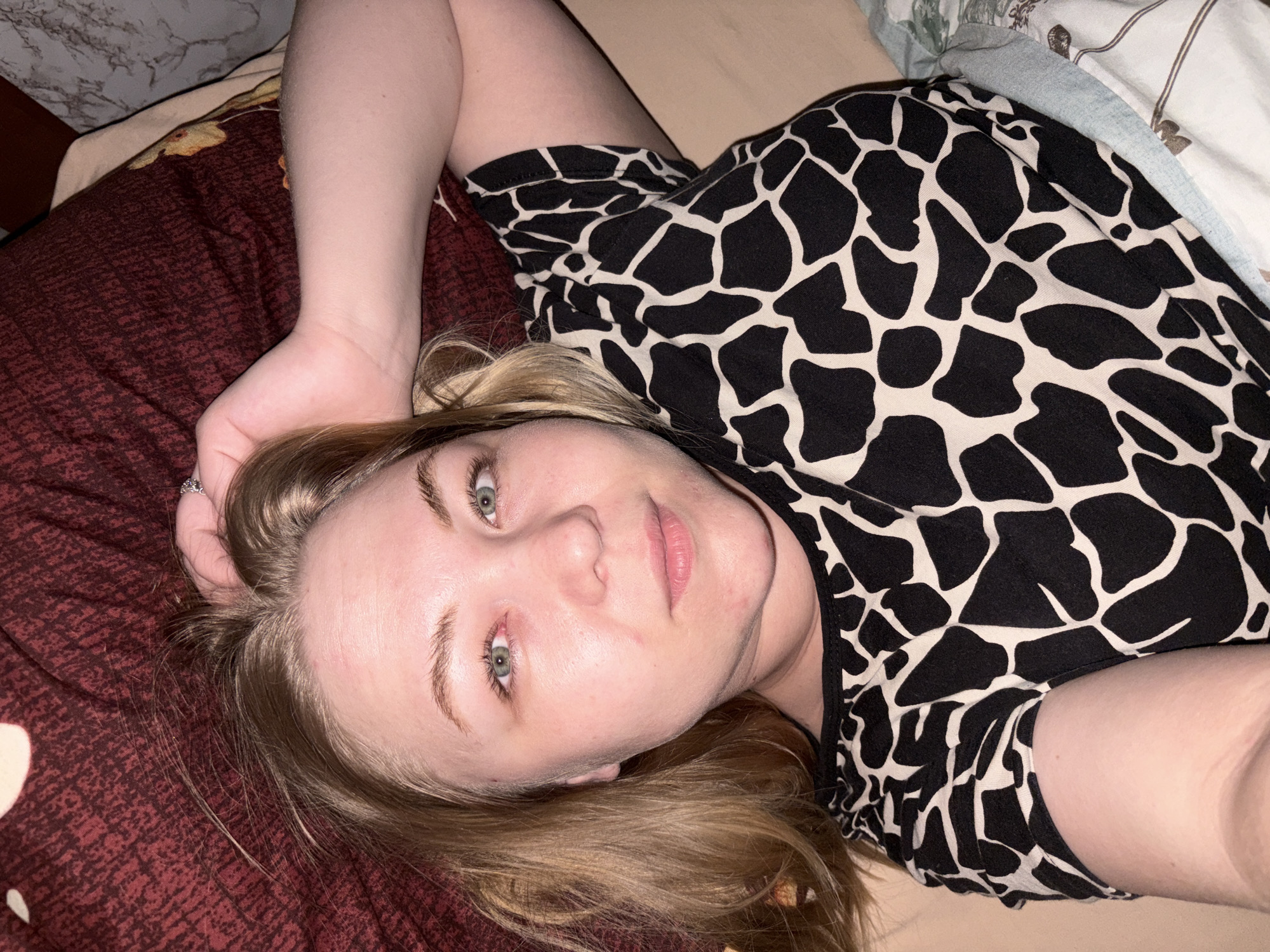 A fair-skinned woman with blonde hair is lying down, looking at the camera. She is wearing a black and white patterned top. She is lying on a bed with a burgundy patterned bedspread and a light-colored blanket with a floral pattern. The location is Noviy, Russia.
