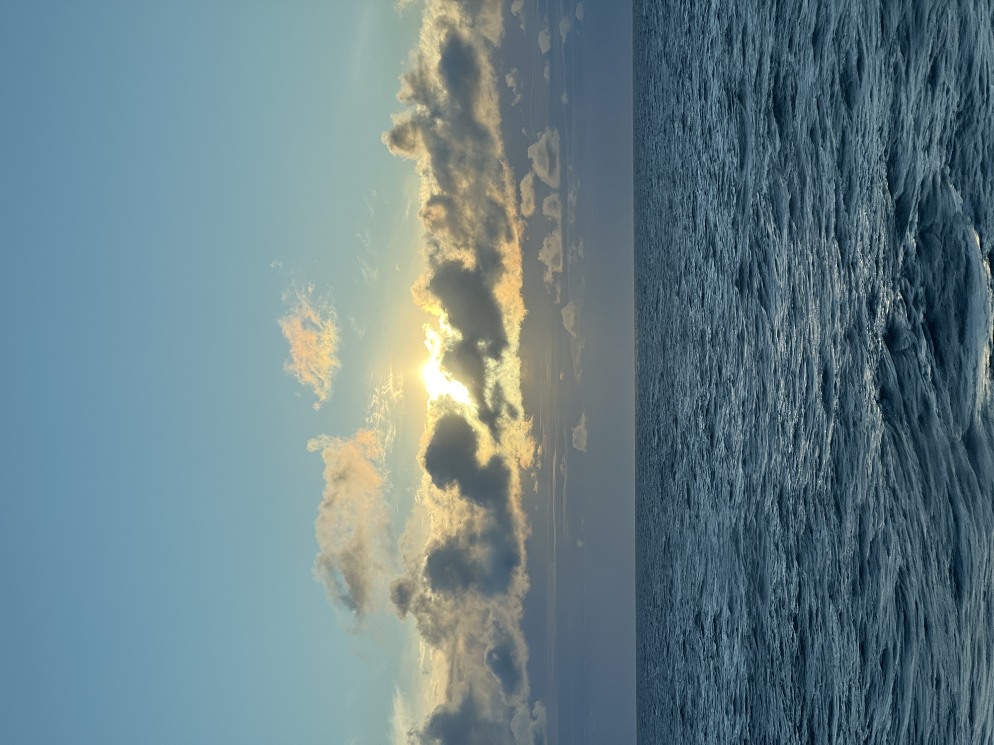 The image depicts a vast expanse of deep blue ocean meeting a sky filled with dramatic clouds during what appears to be sunrise or sunset. The sun's rays pierce through the clouds, casting a warm, golden light on some of them and creating a bright glow. The water's surface is textured with ripples and small waves, reflecting the soft light. The horizon is a clear, straight line dividing the sea and the sky. There are no people or man-made structures visible in the scene. The location is described as Unknown, Poland.