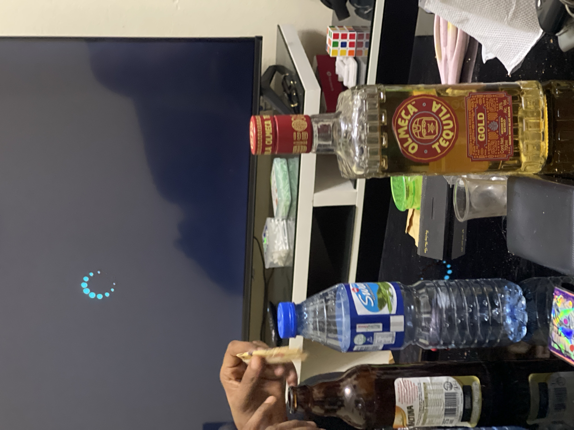 The image shows a cluttered scene indoors. A large flat-screen TV displays a loading animation. In the foreground, a person's hands are visible, holding a small, elongated object. The table surface features an array of items: a bottle of Olmecca Tequila Gold, a bottle of bottled water, and a brown beer bottle. Other assorted items are strewn across the black table. The scene is not identifiable as specifically being in Abuja, Nigeria. No specific details are present that would allow for this determination.
