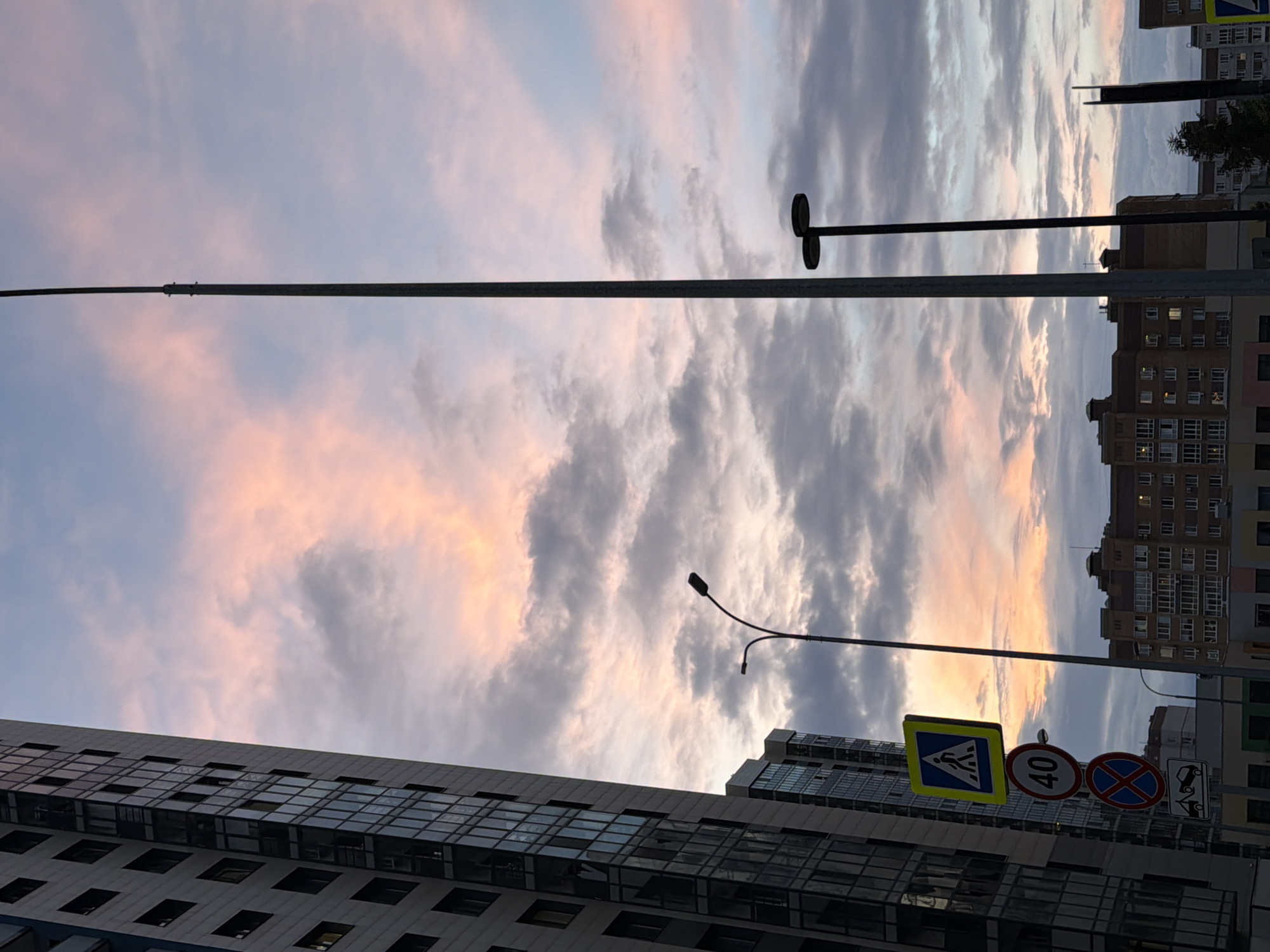 The image depicts an urban scene with a sunset sky. The sky is filled with dramatic clouds, streaked with hues of pink, orange, and grey, indicating either sunrise or sunset. Tall buildings dominate the right side of the frame, characterized by modern architecture with numerous windows. A pedestrian crossing sign, a speed limit sign showing "40", and a "no stopping" sign are visible in the foreground, suggesting a street or intersection. A street lamp with two light fixtures is also present. The perspective is from a low angle, looking up towards the sky and buildings, creating a sense of scale. The overall mood is somewhat dramatic due to the intense sky colors and the imposing urban structures. The image was taken in Kazan, Russia.
