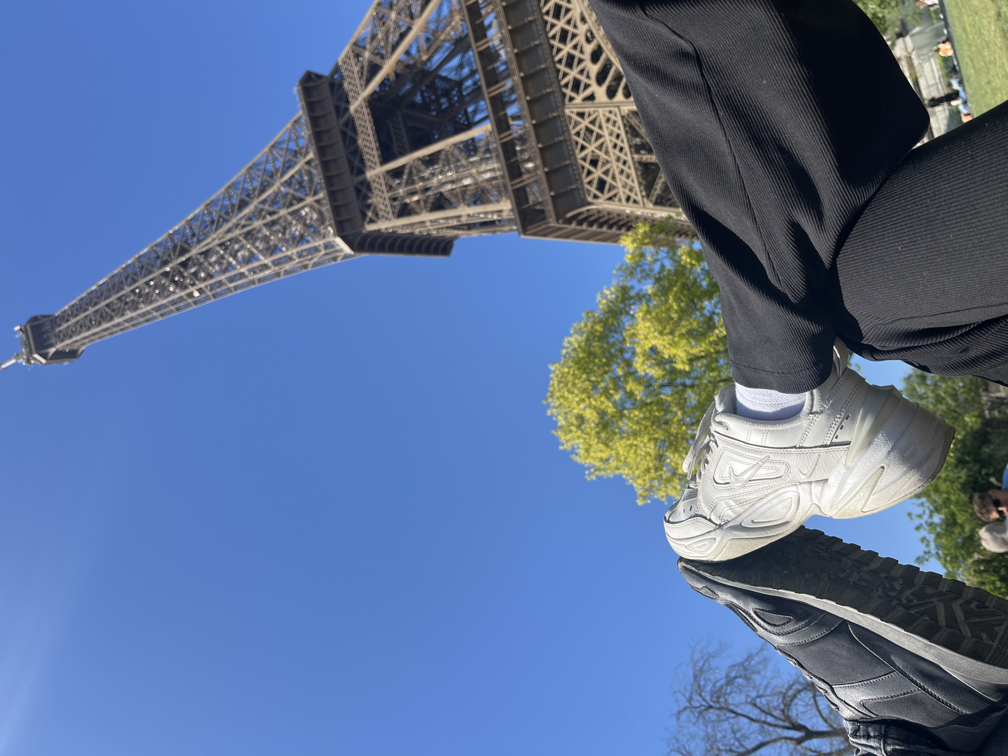 The image shows a low-angle view of the Eiffel Tower in Paris, France, with a clear blue sky visible in the background. Part of a tree with green foliage is visible to the right of the tower. In the foreground, two pairs of legs are visible, presumably belonging to two people. The person on the right is wearing black corduroy pants and a white sneaker. The person on the left is wearing black pants and a dark colored shoe. The Eiffel Tower fills the upper left portion of the image.