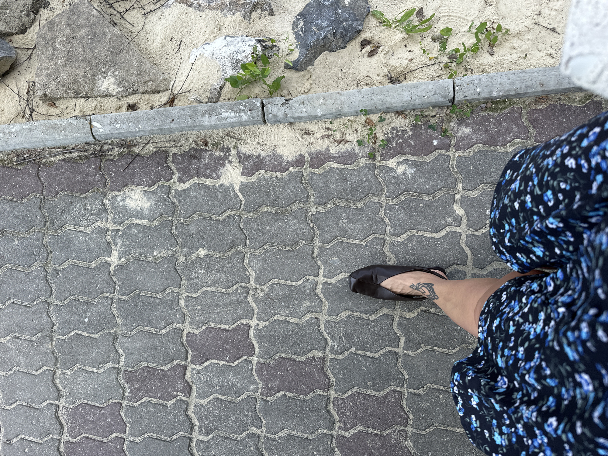 The image shows a top-down view of a person's legs and feet walking on a paved walkway made of interlocking grey and purplish-grey bricks. The walkway is bordered on the right by a sandy area with rocks and sparse vegetation. A damaged concrete curb separates the walkway from the sandy area. The person is wearing a dark blue dress with a floral pattern of light blue and white flowers, and dark, pointed-toe flat shoes. A tattoo is visible on the person's left ankle. The scene suggests an outdoor urban or semi-urban environment, possibly a pathway in Duvankul', Russia.