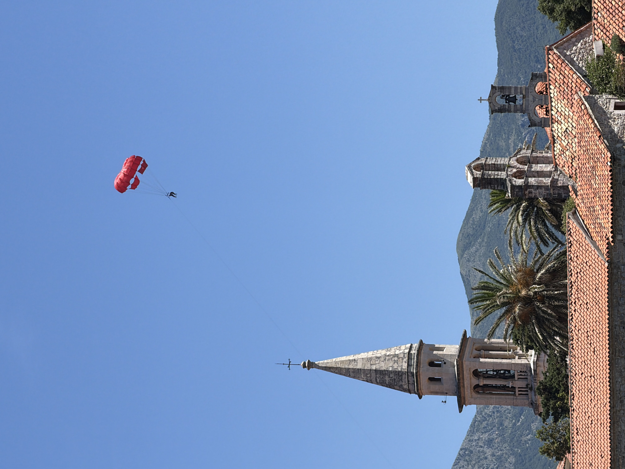 In Budva, Montenegro, a bright red parasail, carrying a single person, drifts across a clear blue sky above the historic Old Town. The parasail is positioned high and to the right of the frame. Below, the architectural landscape of the Old Town unfolds, characterized by terracotta-tiled roofs. Dominating the left side of the lower frame is a tall, stone bell tower, capped with a slender spire, its facade adorned with arched openings. To the right of the bell tower, two smaller stone structures with arched openings and bells are visible, set against a backdrop of lush green palm trees and distant, hazy mountains. The clear, sunny day suggests a recreational activity taking place in a coastal setting.