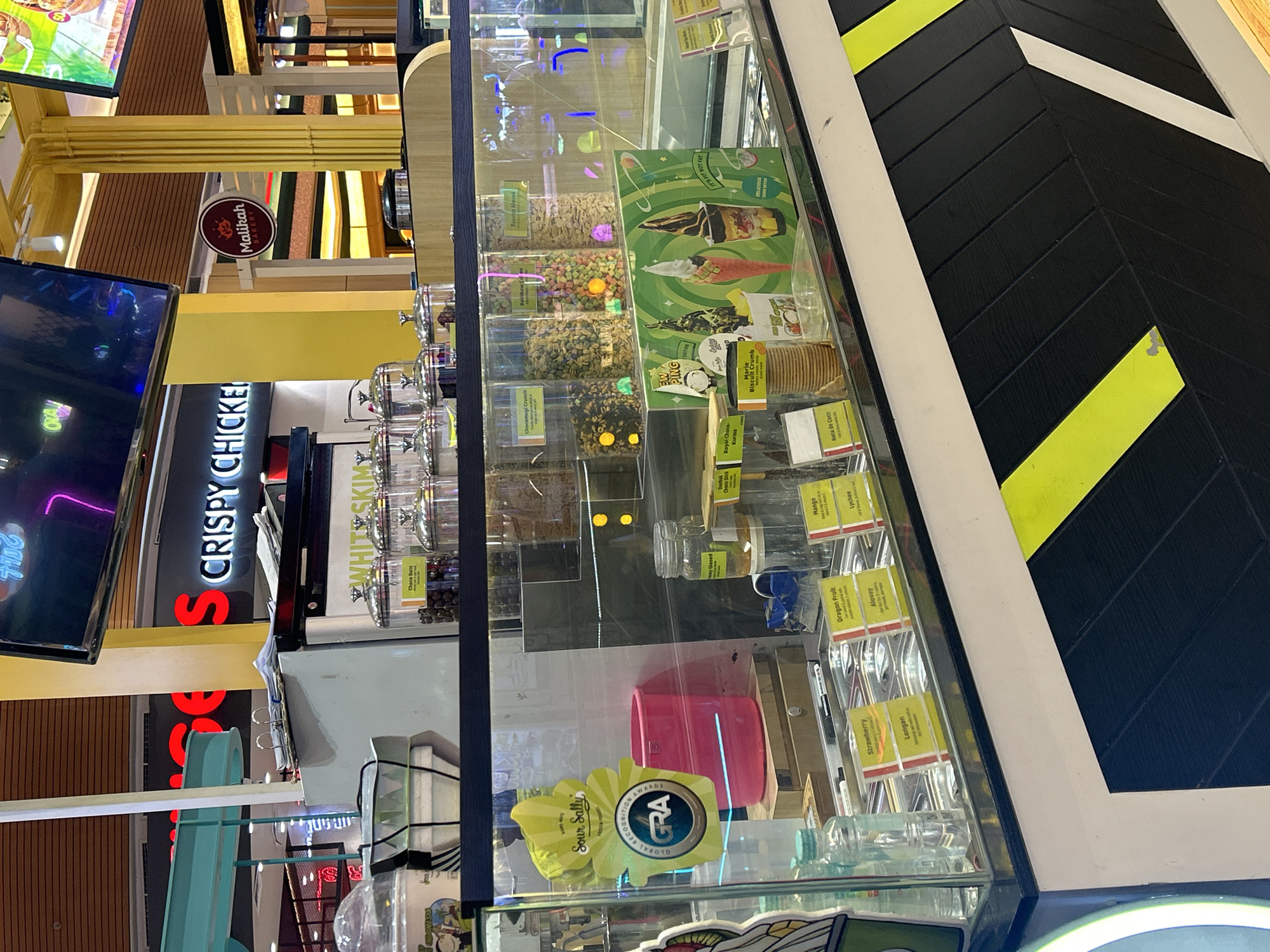 The image shows an ice cream display case inside a food establishment. The display case is illuminated and contains various flavors of ice cream, as well as toppings and cones. There are signs labeling the flavors such as "Strawberry," "Dragon Fruit," "Longan," and "Lychee." In the background, there is a sign for "S Crispy Chicken" and another for "Malikah Bakery." The establishment appears to be located in a mall or a similar indoor shopping area. The overall impression is that of a brightly lit, modern food stall offering frozen treats.