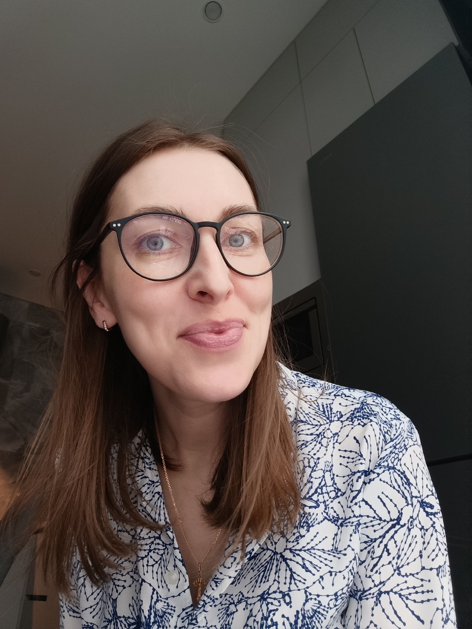 A woman is indoors, making a playful expression with her tongue slightly sticking out. She wears round, black-rimmed glasses and a white shirt with a blue pattern. She has long, brown hair. She is also wearing earrings and a necklace with a pendant. The location is the city of Chaikovsky, Russia. A dark gray wall or panel is in the background.
