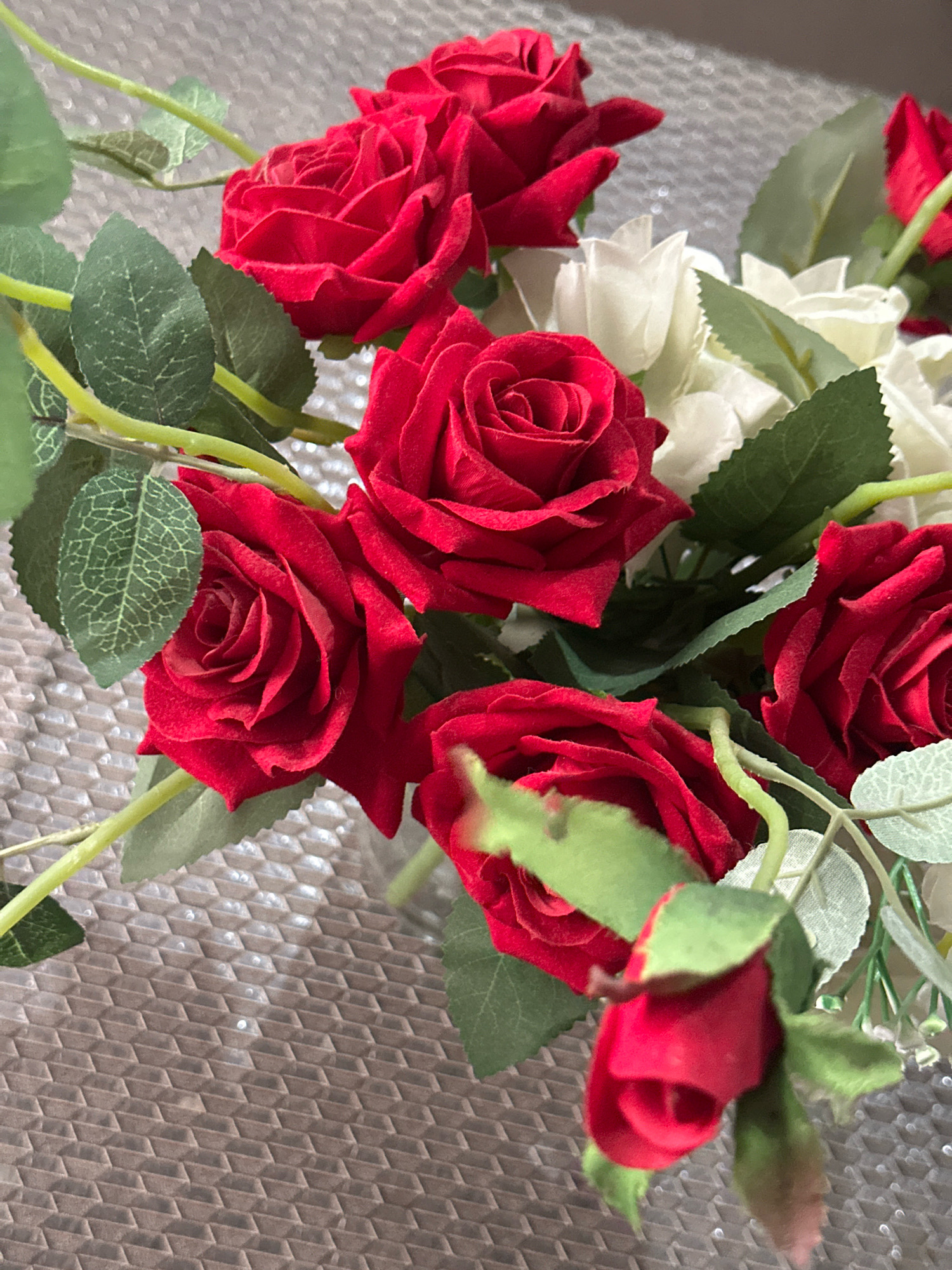 This is a close-up, eye-level shot of a bouquet of artificial red roses and white flowers, with green leaves and stems, resting on a textured, reflective surface. The flowers are arranged in a way that suggests they are part of a larger decorative piece, possibly an arrangement for an event or as a table centerpiece. The lighting is soft, with some highlights on the surface and the flowers, indicating it might be artificial light. There is no visible indication of the location being Taharpur, India; the scene is generic and could be anywhere.