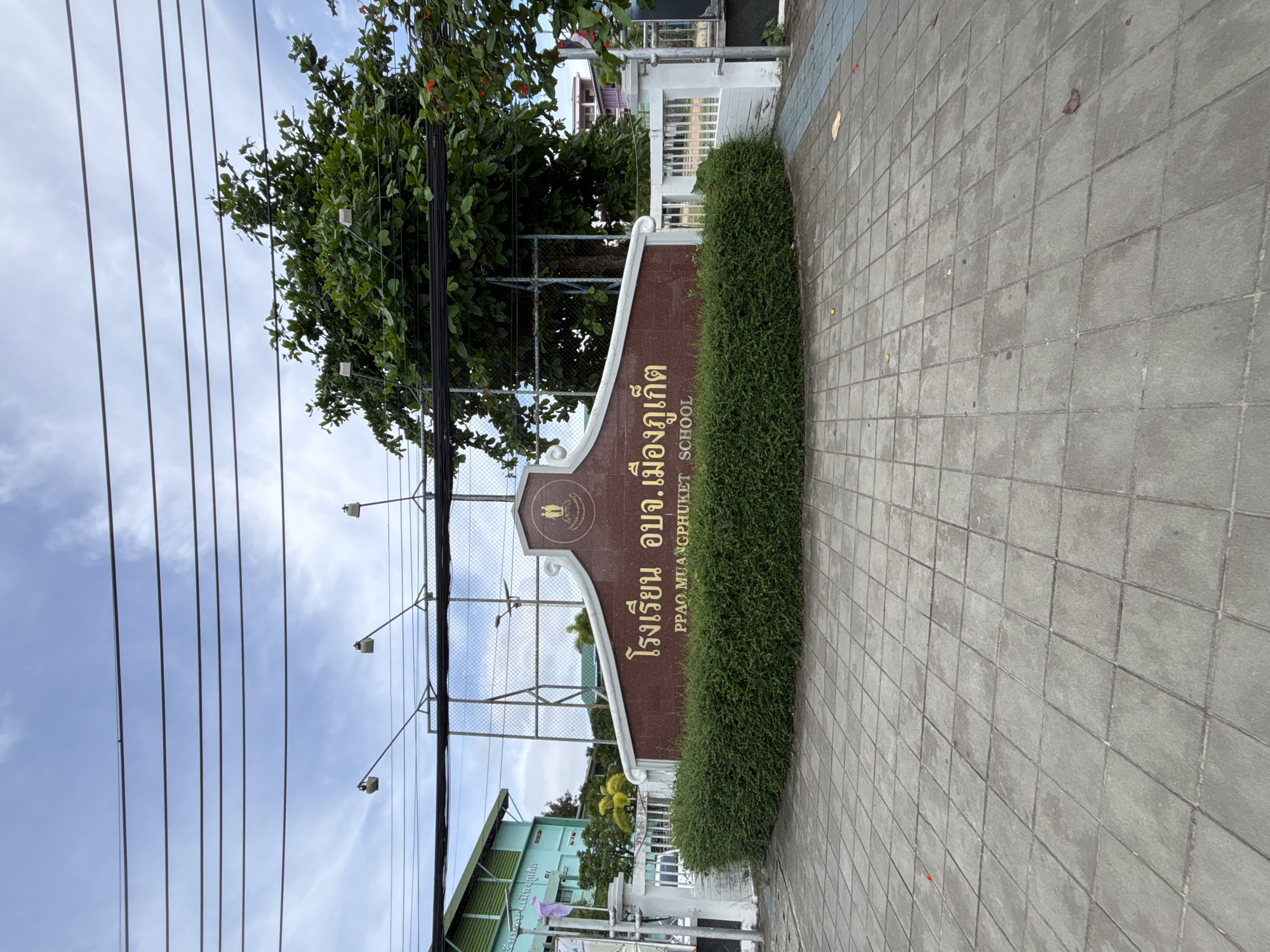 The image depicts the entrance to PPAO Mueang Phuket School in Rawai, Thailand. A large, brown sign with gold lettering is prominently displayed, featuring Thai script at the top and "PPAO MUANGPHUKET SCHOOL" in English below. The sign is bordered by a white decorative element and topped with a school emblem featuring two figures. In front of the sign, a neatly trimmed green hedge forms a semi-circular border. The ground in front of the hedge is paved with large, grey square tiles. Behind the sign, a chain-link fence encloses a sports field, with a large green tree and power lines visible in the background against a cloudy sky. To the left, a portion of a green school building is visible. The overall scene is that of an institutional entrance, implying an educational facility.