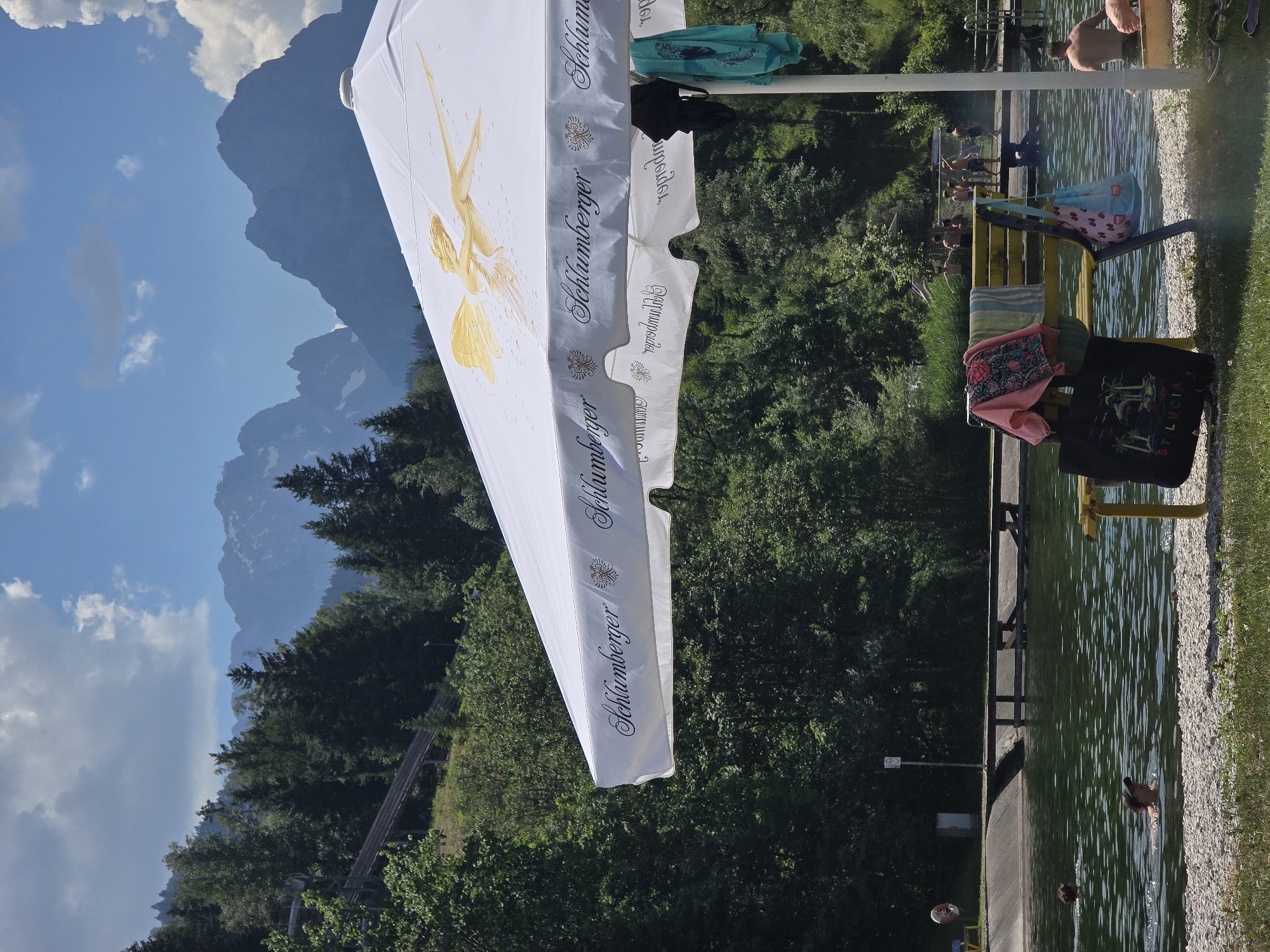 The image depicts a sunny day at an outdoor swimming area, likely a public pool or lake, in Garmisch-Partenkirchen, Germany. The scene is dominated by a large white umbrella with a golden graphic of a winged figure and the "Schlumberger" logo repeated along its edge. The umbrella is partially obscuring a backdrop of lush green trees and imposing mountains, suggesting the setting is nestled within a mountainous landscape. The water of the swimming area is clear and appears to be a vibrant green, with ripples indicating movement. Several people are visible, some enjoying the water and others relaxing by the poolside. On a yellow bench, towels and bags are placed, with one black bag displaying a "ST LUCIA" graphic. The time of day appears to be midday, given the bright sunlight casting shadows and illuminating the scene. The weather is fair and warm, conducive to outdoor recreational activities. The overall mood is relaxed and leisurely, with individuals enjoying a summer day in a scenic natural environment.