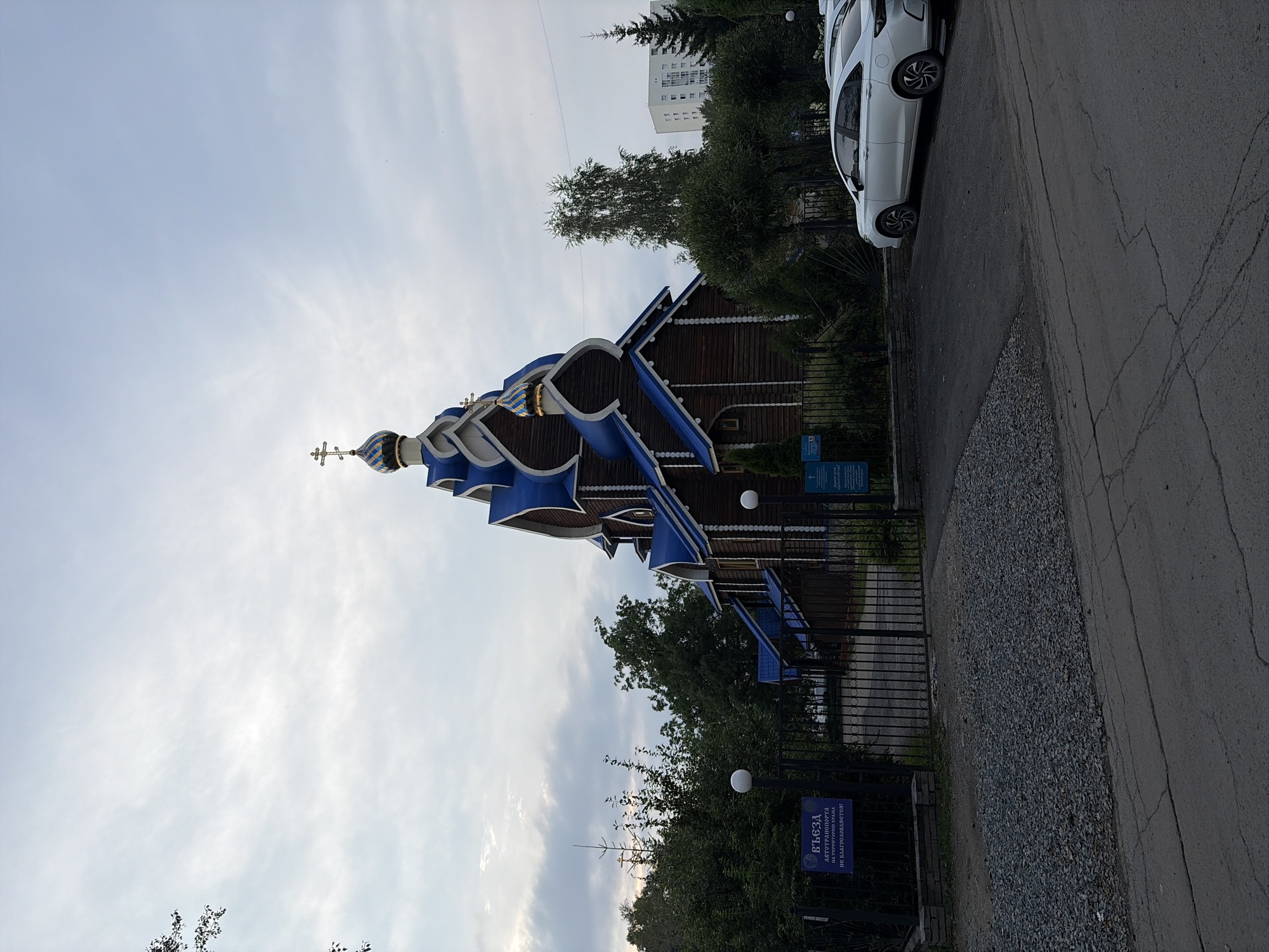 The image shows a wooden Russian Orthodox church with blue onion domes and a golden cross on its tallest dome. The church is located on a street in Yekaterinburg, Russia. In the foreground, there is a black metal gate with a sign that reads "ВЪЕЗДА АВТОТРАНСПОРТА НА ТЕРРИТОРИЮ ХРАМА НЕ БЛАГОСЛАВЛЯЕТСЯ!" which translates to "Entry of vehicles onto the temple territory is not blessed!". A white sedan is parked in front of the church to the right. The sky is cloudy and overcast, suggesting it may be late afternoon or early evening. Trees and bushes surround the church, and a modern apartment building can be seen in the background. The ground is a mix of gravel and asphalt.