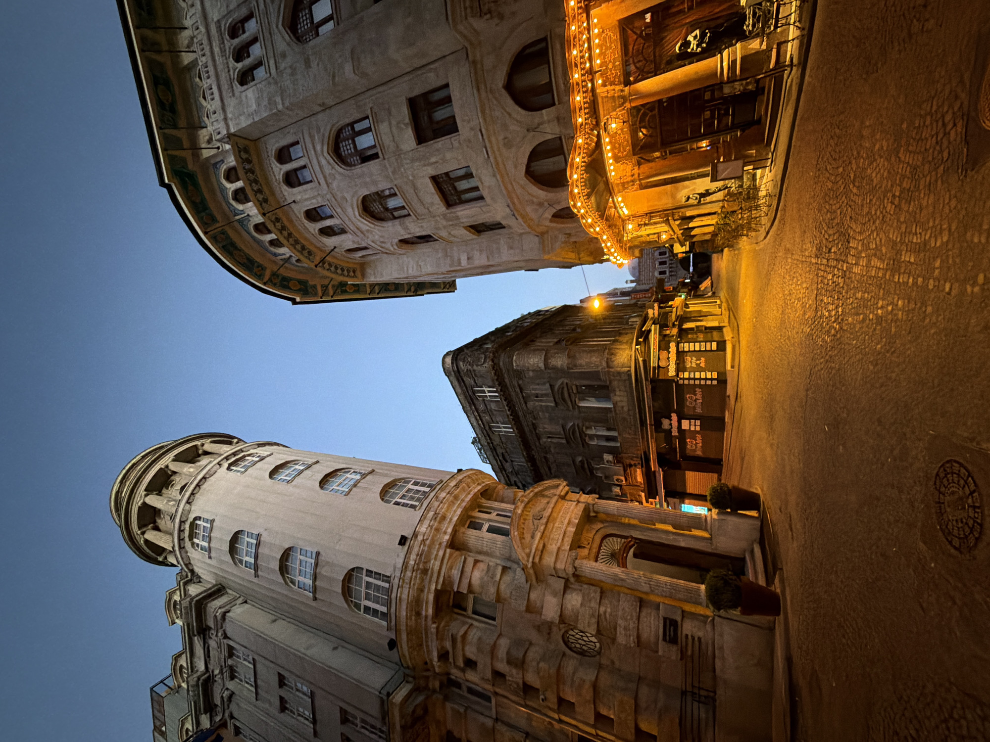 The image captures a street scene in Istanbul, Turkey, at dusk. The perspective is low, looking upwards at two prominent, older buildings flanking a narrow, cobblestone street. The building on the left is a rounded, multi-story structure with classical architectural elements, including columns and arched windows. The building on the right is also multi-story with ornate, arched windows and decorative friezes. Its ground floor features a brightly lit entrance with strings of warm-toned lights, suggesting it is a restaurant or bar with outdoor seating. The street is paved with cobblestones, and the warm glow from the building's lights illuminates the pavement. Further down the street, another older, more angular building is visible, and beyond that, a dome structure hints at more traditional Ottoman architecture in the distance. The sky above is a clear, deep blue, indicating the transition from daylight to night. No people are visible in the frame. The overall impression is one of historic urban architecture illuminated by artificial light in the twilight.