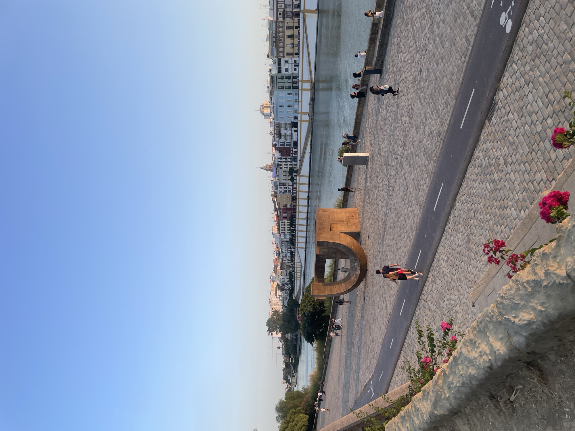 The image shows a riverside promenade in Seville, Spain, during what appears to be late afternoon or early evening. The Guadalquivir River flows to the right, with a distinct line of colorful buildings lining the opposite bank. In the foreground, a paved area with cobblestone patterns transitions into a dedicated bike lane with white markings. A large, abstract metal sculpture, forming a curved shape resembling a "U," is prominently positioned on the promenade.
Several individuals are visible, engaged in various activities. Some are walking along the promenade, while others are seated on a low wall near the river. Two people are walking together on the bike lane, a man and a woman. Further down the promenade, other pedestrians can be seen in groups and individually. The sky is clear and a soft blue, indicating favorable weather. Lush green trees are present along the riverbank. Pink bougainvillea flowers add a touch of color to the lower left and lower right corners of the image, cascading over a stone wall. The overall atmosphere is relaxed and pleasant, suggesting a popular spot for recreation and sightseeing in Seville.