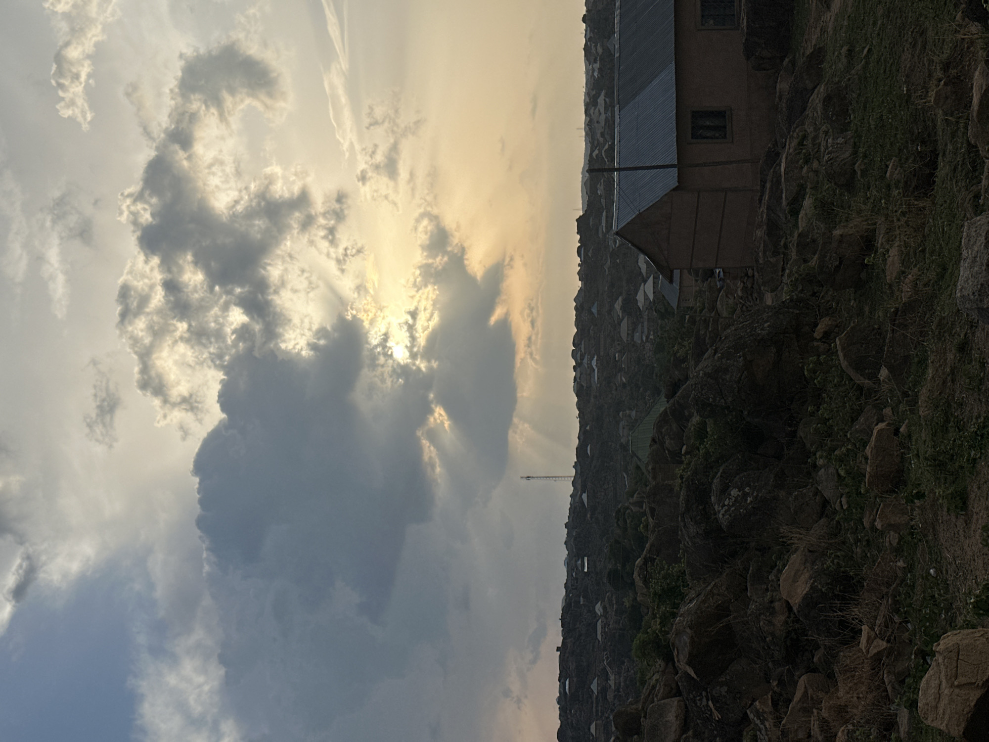 The image depicts a landscape scene in Jos, Nigeria, under a cloudy sky with the sun partially obscured. The foreground is dominated by rocky terrain and sparse vegetation. In the midground, buildings are clustered together, some with metallic roofs, giving the impression of a dense settlement on elevated ground. A communication tower is visible amidst the buildings. The sky is filled with grey and white clouds, with sun rays breaking through in places.