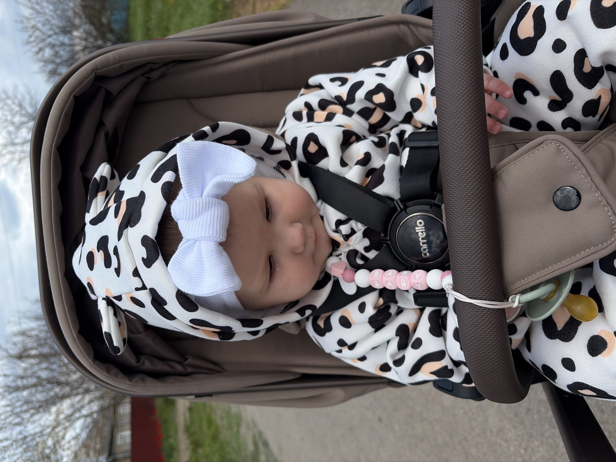 An overhead view of an infant lying in a stroller, rotated 90 degrees counter-clockwise. The baby's head is positioned towards the bottom-left of the frame, with its body extending towards the top-right. The infant appears to be asleep, with eyes closed, and wears a white hooded jumpsuit featuring black and light peach leopard or cow print spots, complete with small ears on the hood. A white ribbed bow headband is on the baby's head.
The baby is nestled in a brown/tan stroller with a visible safety harness and padded interior. A dark brown, textured stroller bar runs diagonally across the upper right of the image. Attached to the baby's clothing is a pacifier clip made of pink and white beads that spell out "BABY". A light green pacifier with a yellow nipple is secured to the stroller bar with a white string. The brand "carrello" is visible on a black buckle mechanism near the baby's chest.
The background, mostly out of focus, shows an overcast sky, bare tree branches, and green ground cover, suggesting an outdoor setting during daytime, likely in spring or autumn. The scene depicts a calm moment in Novopokrovskaya, Russia.