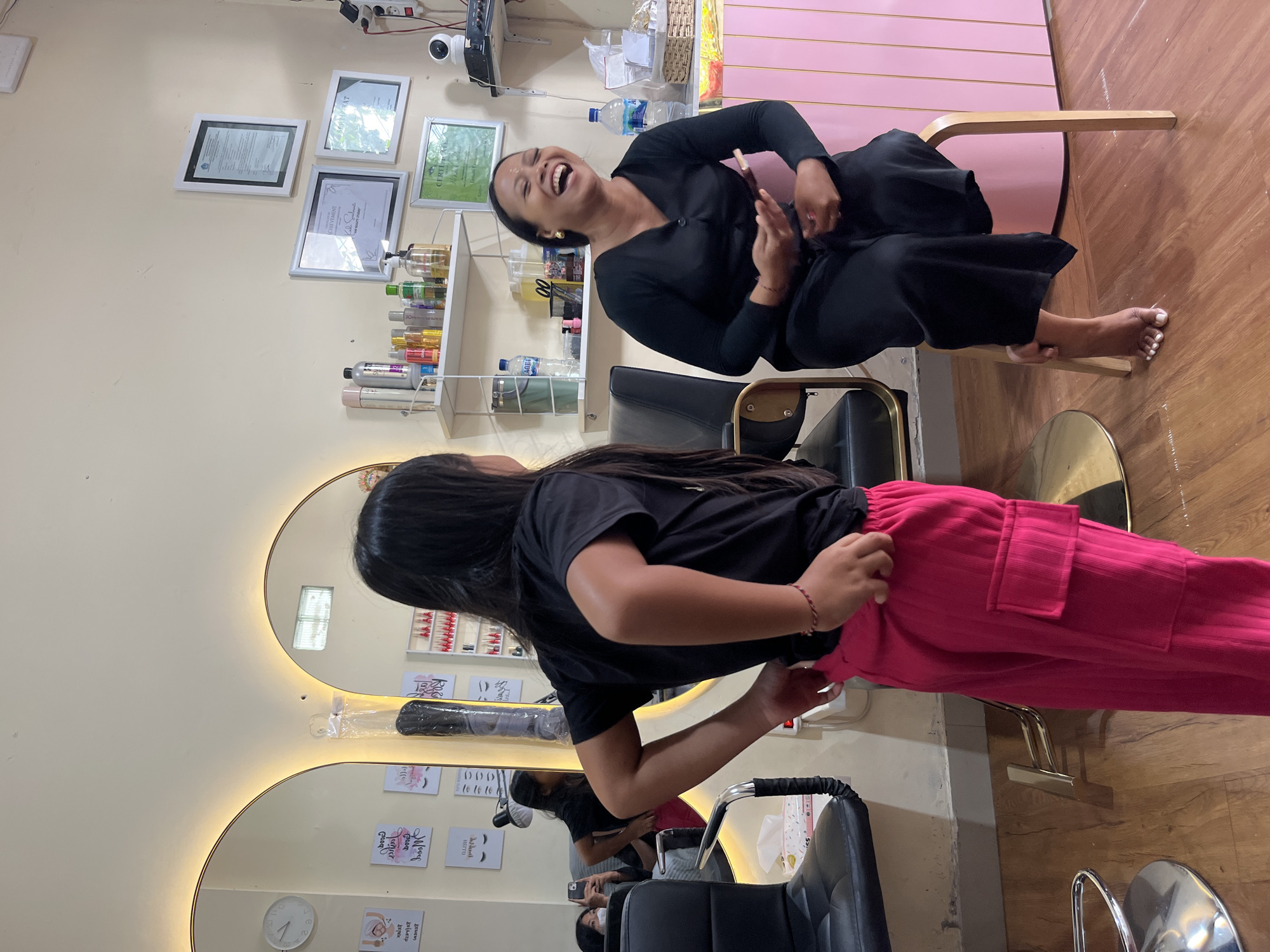 In a beauty salon located in North Kuta, Indonesia, a young girl with long black hair, wearing a black t-shirt and bright pink pants, stands with her back to the camera, facing a woman who is seated. The woman, dressed in a black outfit, is laughing heartily while holding a small rectangular object, possibly a phone or a tablet. She is sitting on a light-colored stool in front of a pink cabinet. The salon appears to be well-lit, with warm lighting coming from behind two large mirrors on the left side of the image. Several framed certificates and shelves stocked with beauty products are visible on the wall. A black salon chair is positioned to the left of the frame, and the floor is made of polished wood. The overall atmosphere suggests a moment of shared amusement within the salon environment.