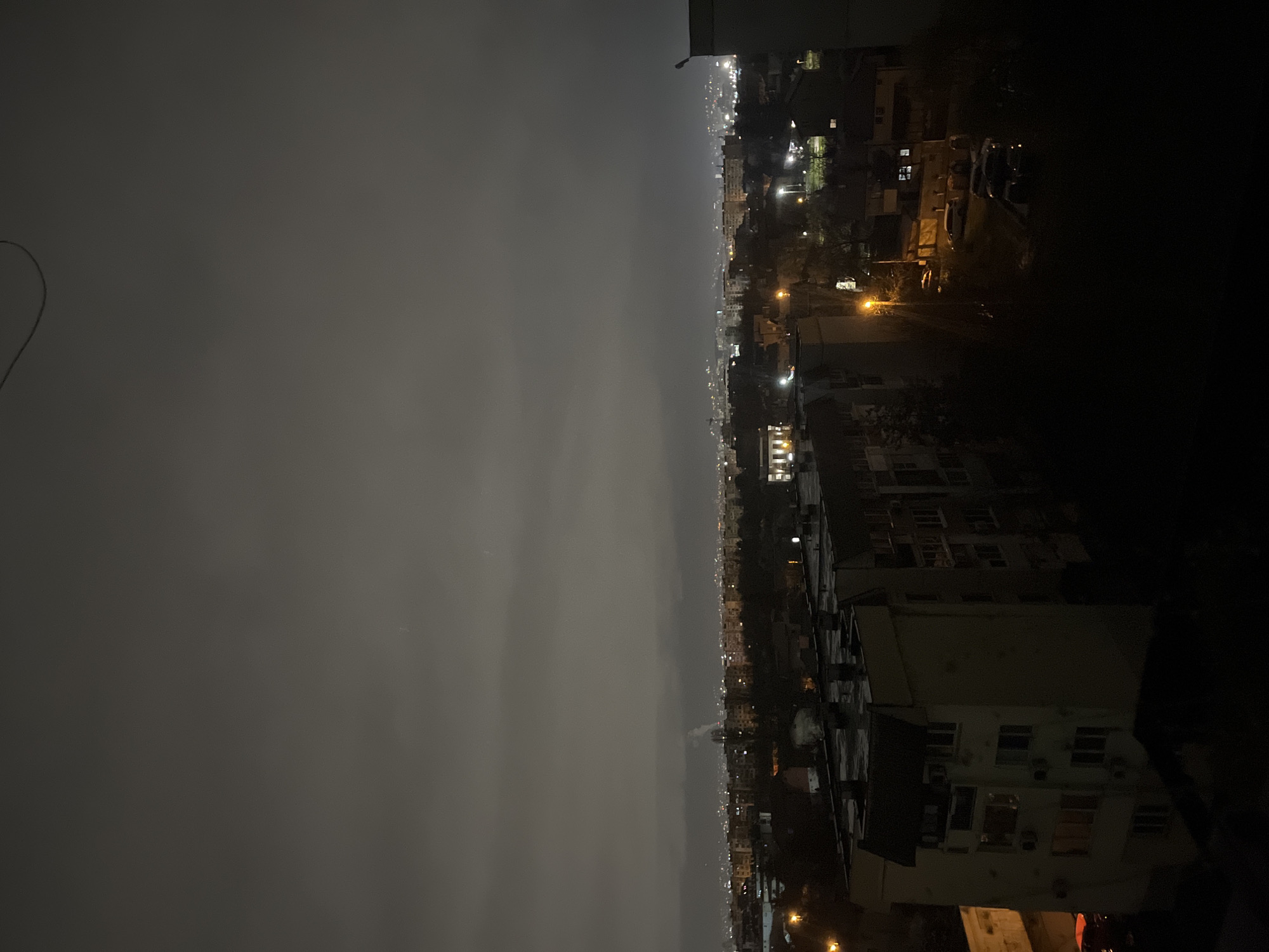 The image is a nighttime cityscape, possibly taken from a high vantage point. On the right, there are lit buildings, with visible windows and some street lights. On the left, the sky is overcast. The location might be Sofia, Bulgaria given the urban structures and general lighting. However, specific details to confirm the precise location are limited.