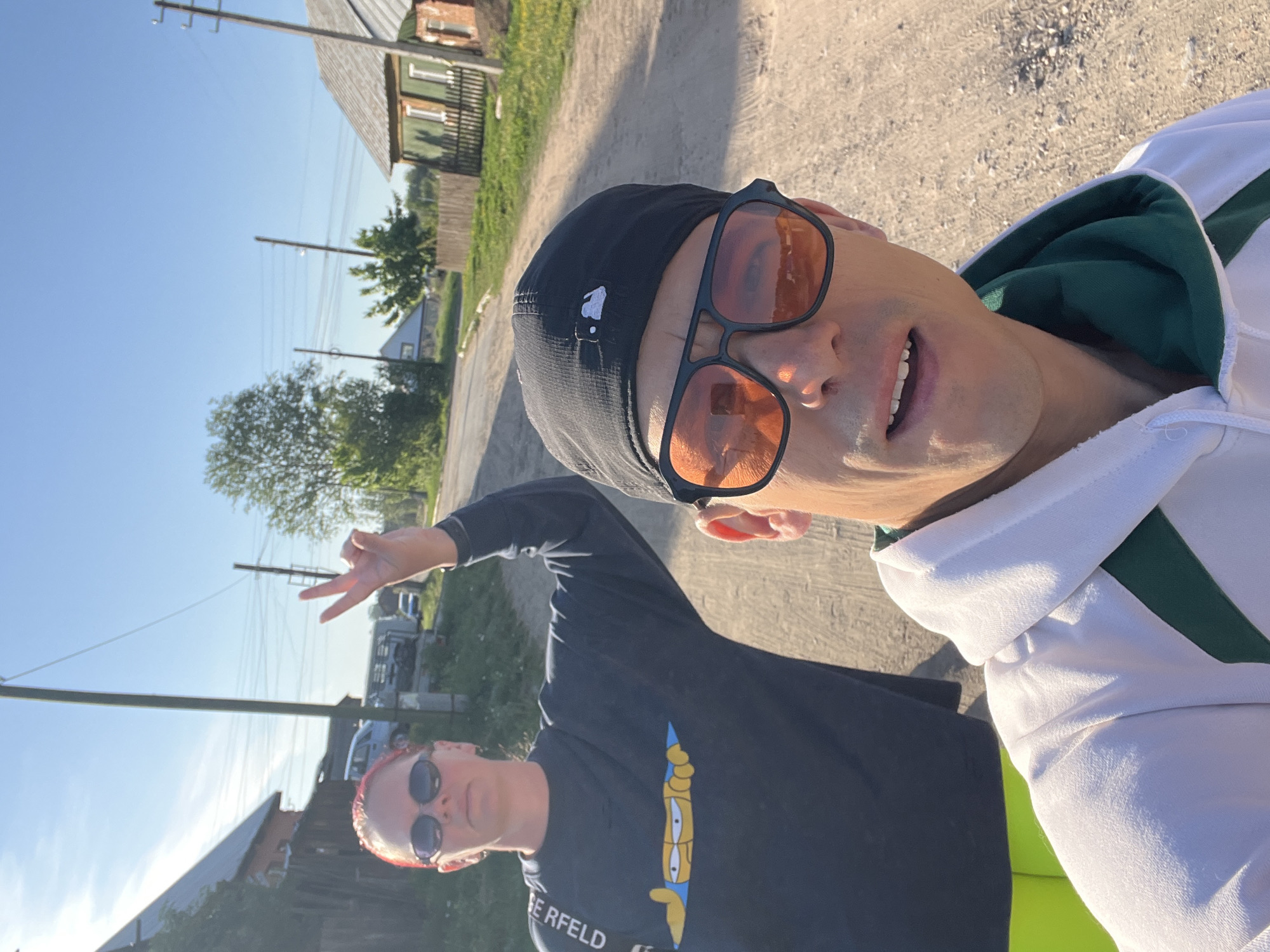 A photo taken on a bright, sunny day in Chaikovsky, Russia, shows two people on what appears to be a dirt road or path. The person taking the photo is an adult male wearing a white hoodie with green trim, a black baseball cap, and orange-tinted sunglasses. His mouth is slightly open as if speaking or posing. In the foreground and to the side of the photographer is another adult, dressed in a black shirt with an illustration from The Simpsons on the front, and sunglasses. This second adult is making a peace sign with their fingers. The background shows a rural scene with houses, trees, and power lines. The overall setting indicates a relatively undeveloped or countryside area within Chaikovsky.
