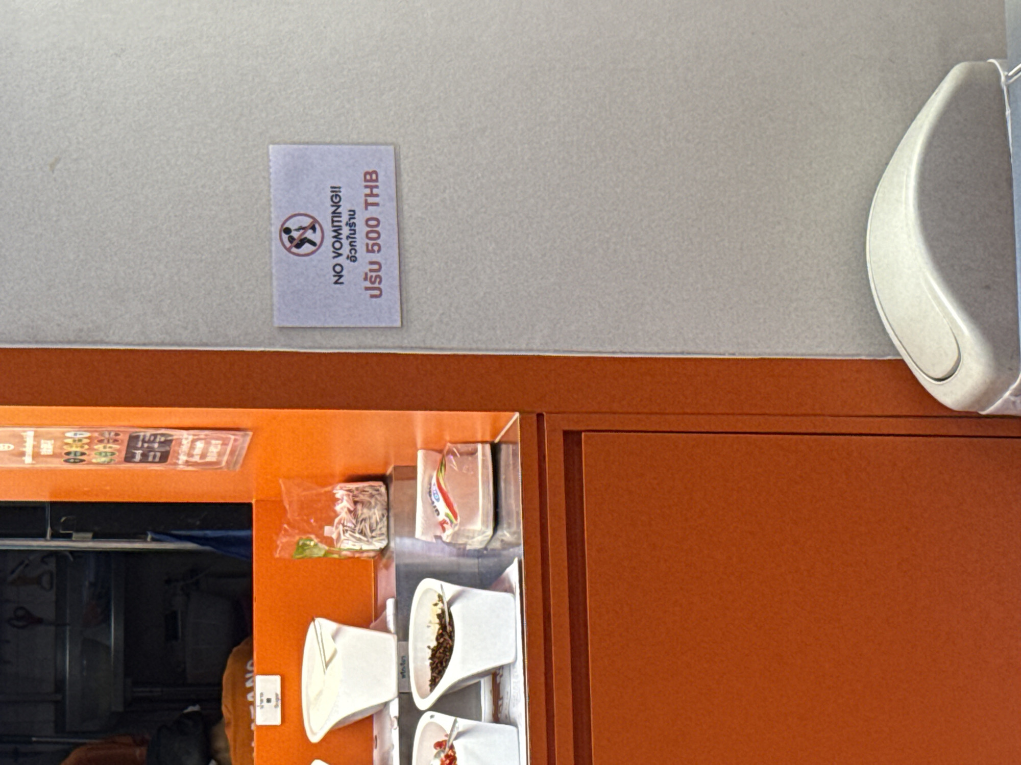 The image shows the upper portion of an orange cabinet/counter setup inside an establishment in Bangkok, Thailand. A white wall is above the cabinet. A sign is adhered to the wall reading "NO VOMITING!!", with an image of a person vomiting, and below it in Thai, a sentence that translates to "Fine 500 THB" (Thai Baht). A white hand sanitizer dispenser is attached to the wall near the right side of the frame. On the counter, a variety of food items and condiments are arranged, including small containers of what appears to be toppings, packets of condiments and other food items.