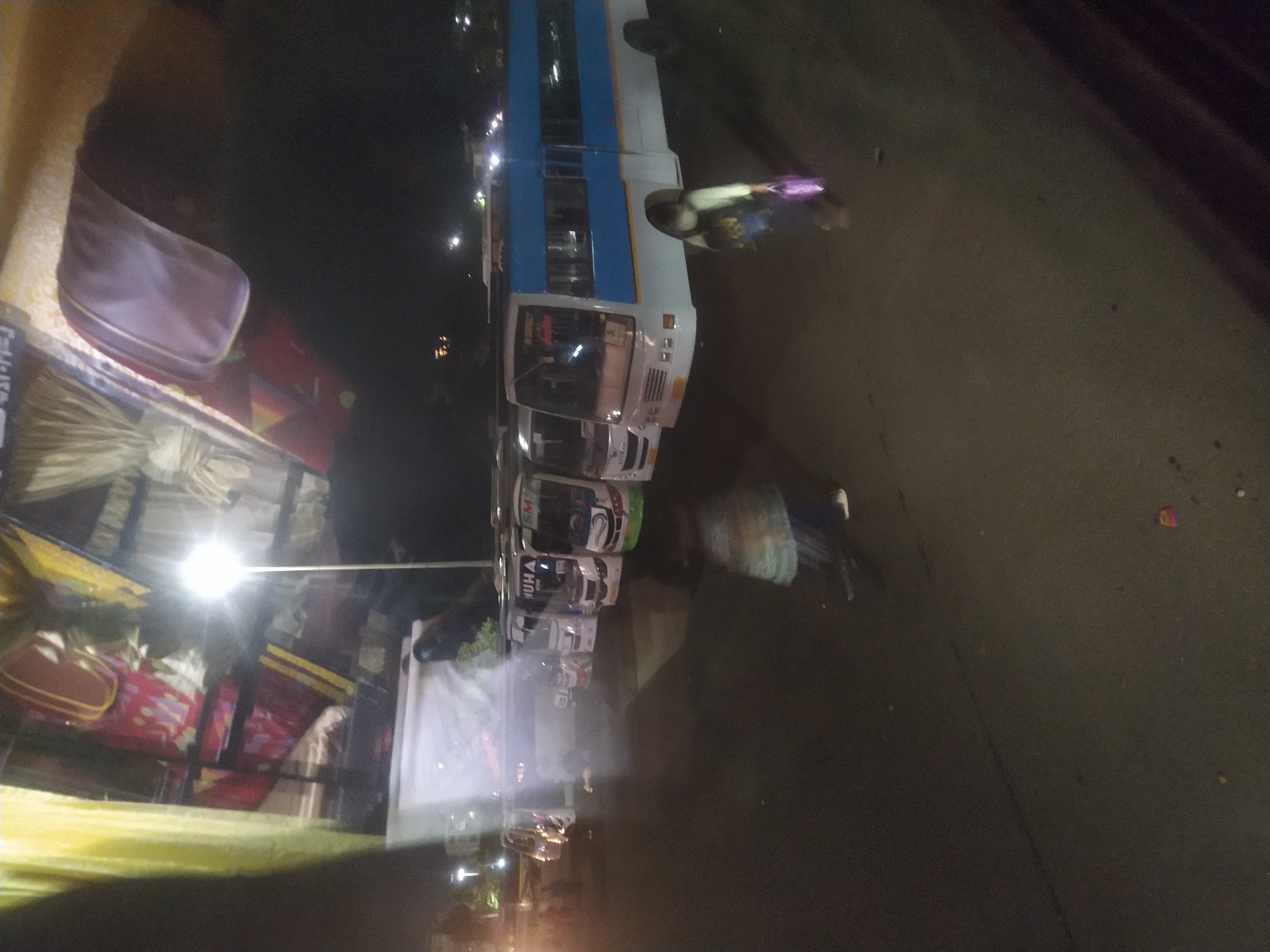 At night in Margao, India, several buses are parked along a street. One bus is white with a blue stripe. People are near the bus, and objects, including luggage, are visible in the background near a light.