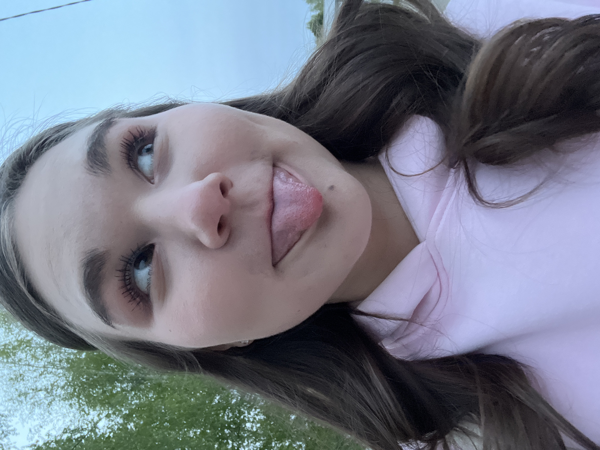 A young woman with long brown hair and wearing a light pink hooded sweatshirt sticks her tongue out. She is looking upward and to the left of the frame. The background includes green foliage and a light blue sky. The location is not discernible from the image.