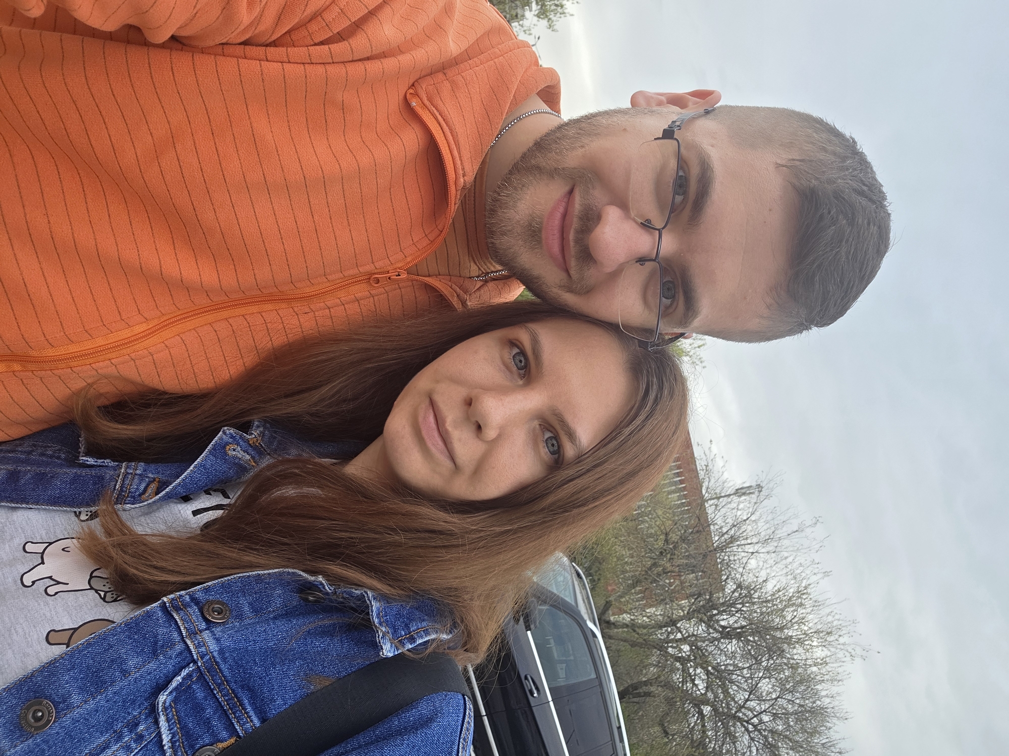 A selfie of a man and a woman, seemingly a couple, taken outdoors on an overcast day. The man is wearing an orange striped zip-up jacket and glasses. He has short hair and a beard. The woman has long brown hair, a denim jacket over a white t-shirt with cartoon animals, and appears to be wearing a seatbelt. They are standing next to a dark-colored vehicle with a building and bare trees in the background. It's not possible to pinpoint the exact location in Plovdiv without additional information.
