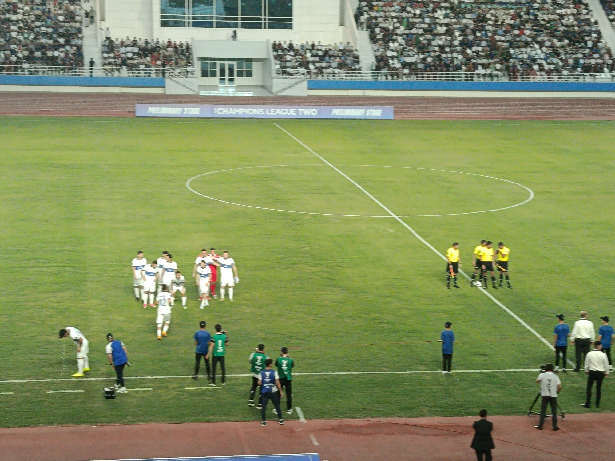 This is a wide, high-angle shot of a soccer match in progress at the Arkadag Stadium in Arkadag City, Turkmenistan. The stadium is filled with spectators in the stands, mostly wearing white and dark clothing. A large, vibrant green soccer field dominates the frame, marked with white lines for the game. In the foreground, a group of players in white and blue uniforms are gathered near the center line. On the right side of the field, a referee in a yellow shirt and black shorts stands with other officials. A cameraman is positioned near the sideline, filming the match. In the background, a large electronic billboard displays "PRELIMINARY STAGE CHAMPIONS LEAGUE TWO PRELIMINARY STAGE." The overall scene depicts a professional sporting event with a focus on the field and the participants.