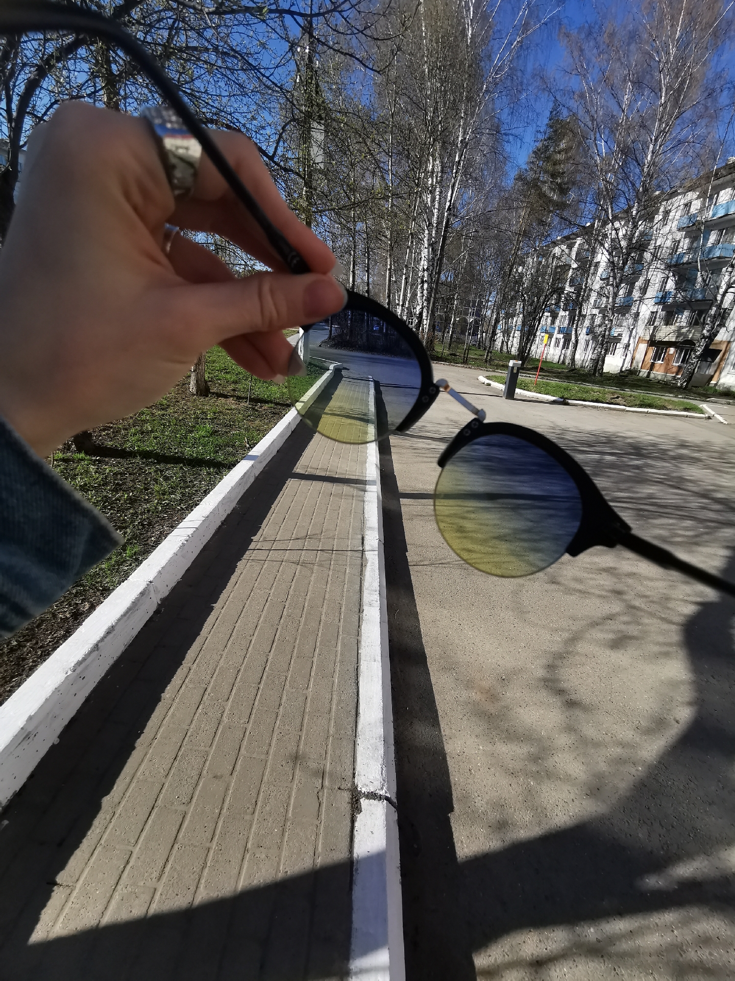 A close-up shot shows a hand holding a pair of sunglasses against an outdoor backdrop. The sunglasses have round, dark frames and lenses with a blue-to-yellow gradient. The hand, adorned with a silver ring, is positioned in the left foreground. The background consists of a paved path bordered by a white painted curb, leading to an apartment complex and bare trees under a clear blue sky. Shadows from the trees and the sunglasses fall across the path.