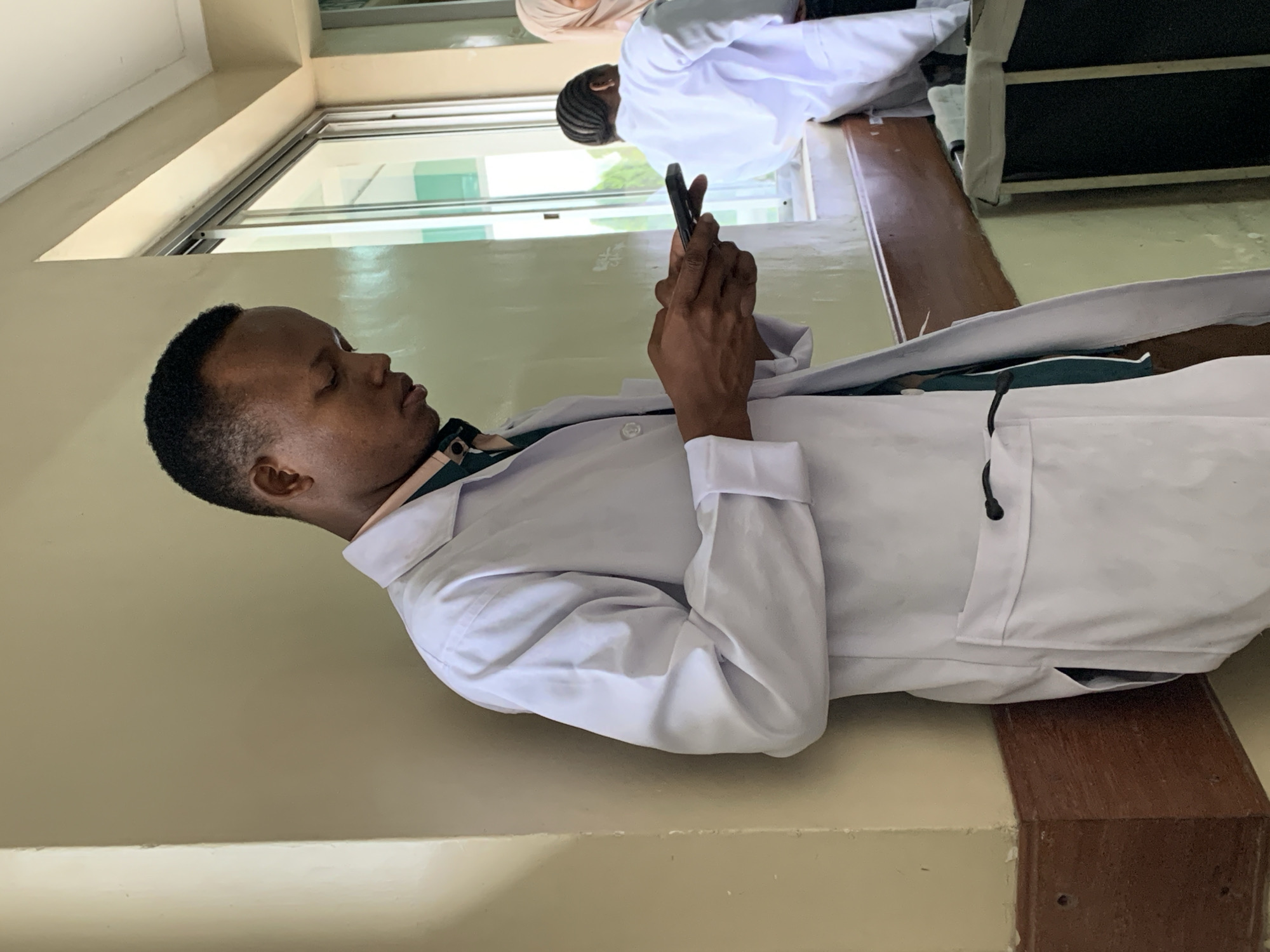 A man in a white lab coat is looking at his cell phone while leaning against a wall. Another person wearing a white lab coat and a hijab is in the background. The location appears to be indoors. The city of Dar es-Salaam, Tanzania cannot be verified from the image.