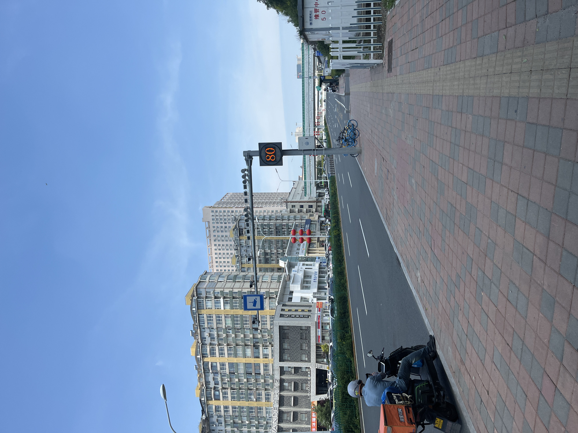 This is a street scene in Changchun City, China, on a clear day. In the foreground, a person sits on a black scooter with an orange delivery box attached to the back. The scooter is parked on a brick sidewalk next to a multi-lane road. The road has white lane markings and several cars are parked along the side. On the left side of the road, tall, multi-story apartment buildings with yellow and gray facades dominate the skyline. A street lamp extends over the road from the left. Further down the road, a digital speed limit sign displays "80". Several blue shared bicycles are parked near the speed limit sign. Across the road, a modern pedestrian overpass with a green roof crosses the street. In the distance, more buildings are visible. On the right side of the image, a white picket fence encloses an area with a small white structure labeled with Chinese characters and the number "50". The sky is a clear blue with a few wispy clouds.