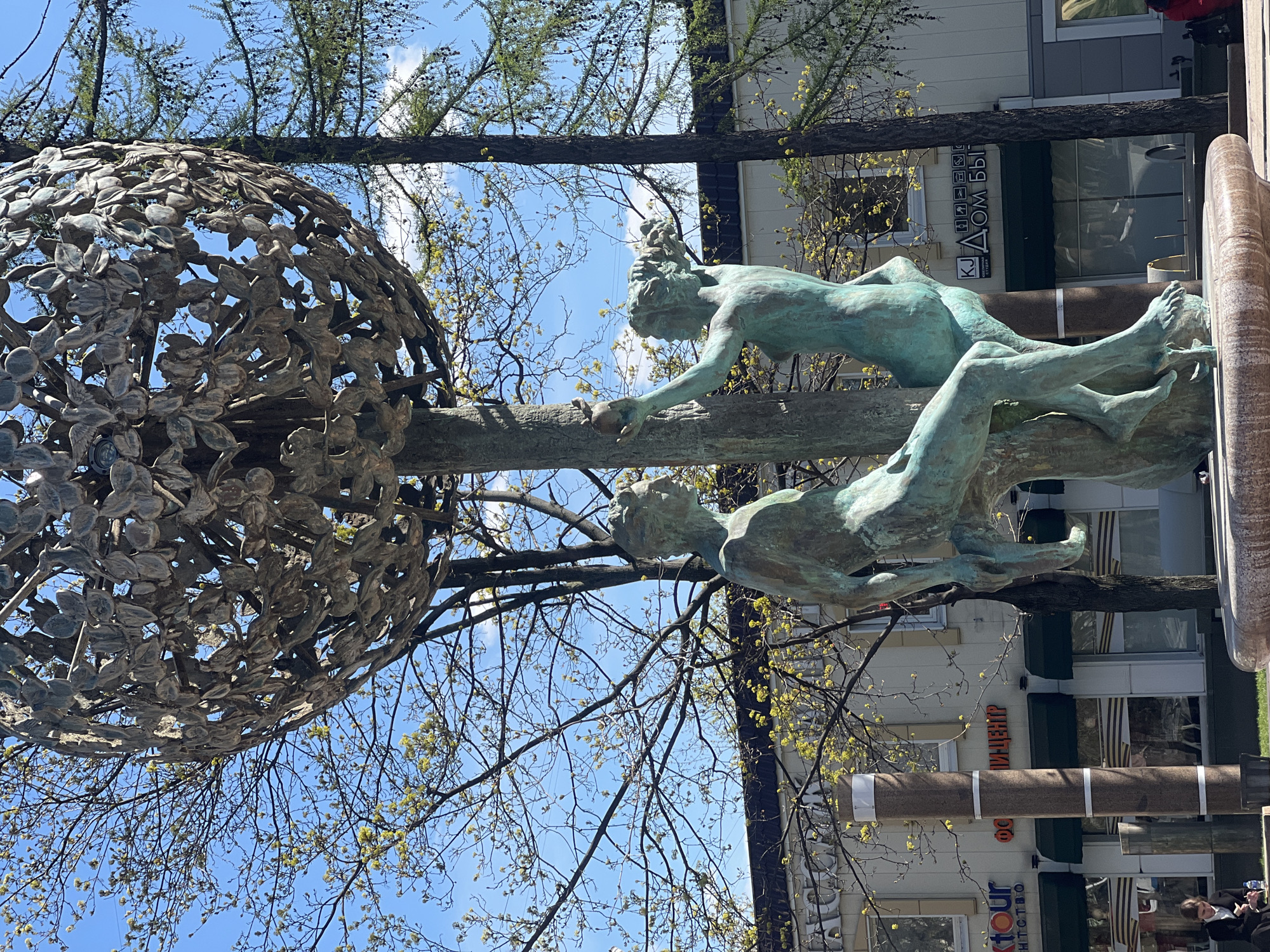 The image depicts a bronze statue of two reclining figures reaching for fruit on a tree. The statue is in a public space in Moscow, Russia. The background includes a building with signs in Russian and the tops of bare trees against a blue sky.