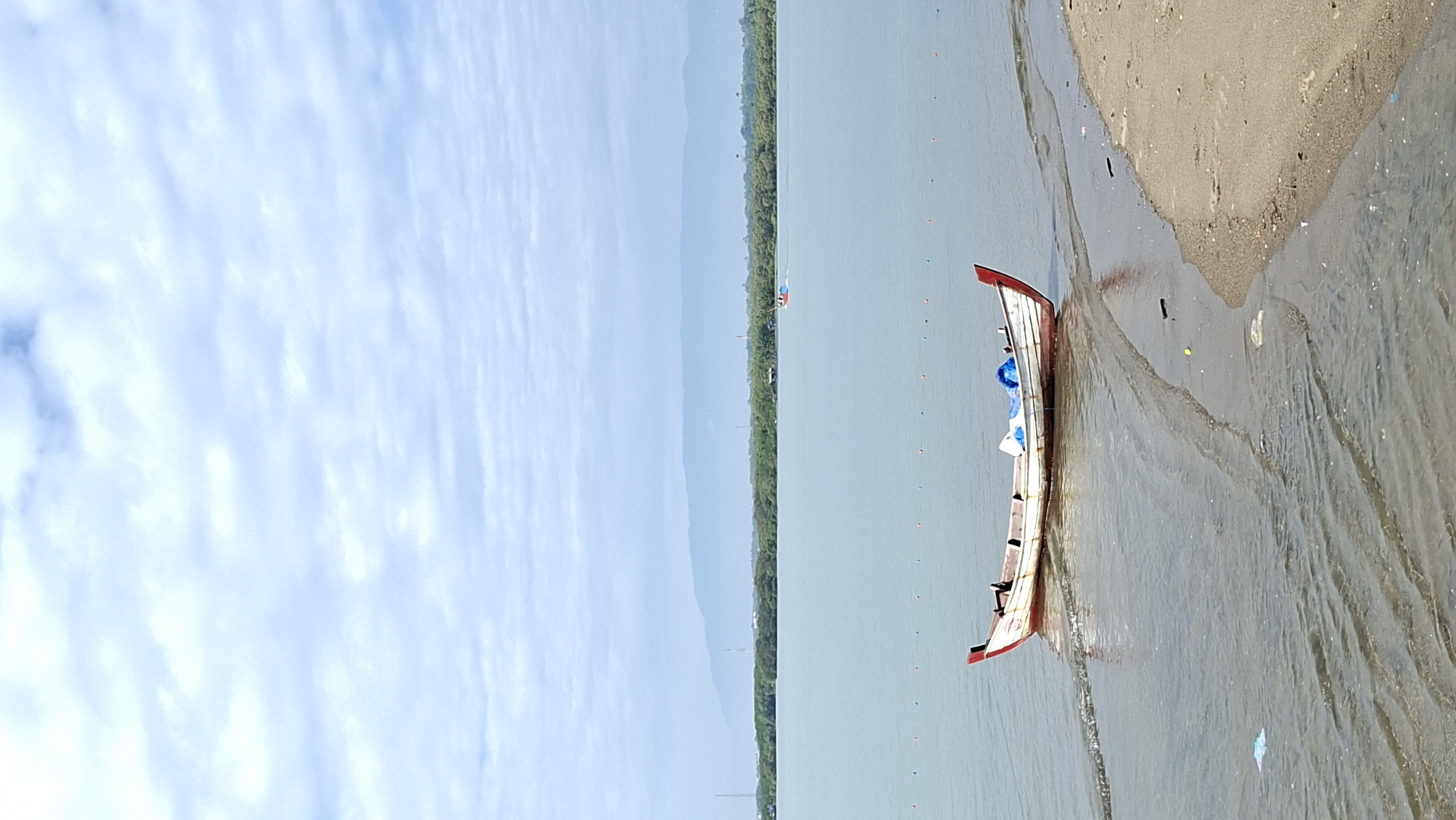 A white and red wooden boat is partially submerged in shallow water near a sandy shore. The boat is positioned diagonally, with its bow facing towards the left and its stern towards the right. It appears to be empty, with some blue material visible inside. The water is calm, with gentle ripples and a sandy seabed visible beneath the surface in some areas. The shoreline is a mix of sand and some debris. In the background, a calm body of water stretches out, leading to a distant tree line and hazy mountains under a cloudy sky. There are buoys visible in the water, indicating a boundary. The overall scene depicts a peaceful coastal environment. While the image is not specific to a particular landmark in Lhokseumawe, the presence of boats and the general coastal landscape are common features of the city's geography, which is situated on the northern coast of Aceh.