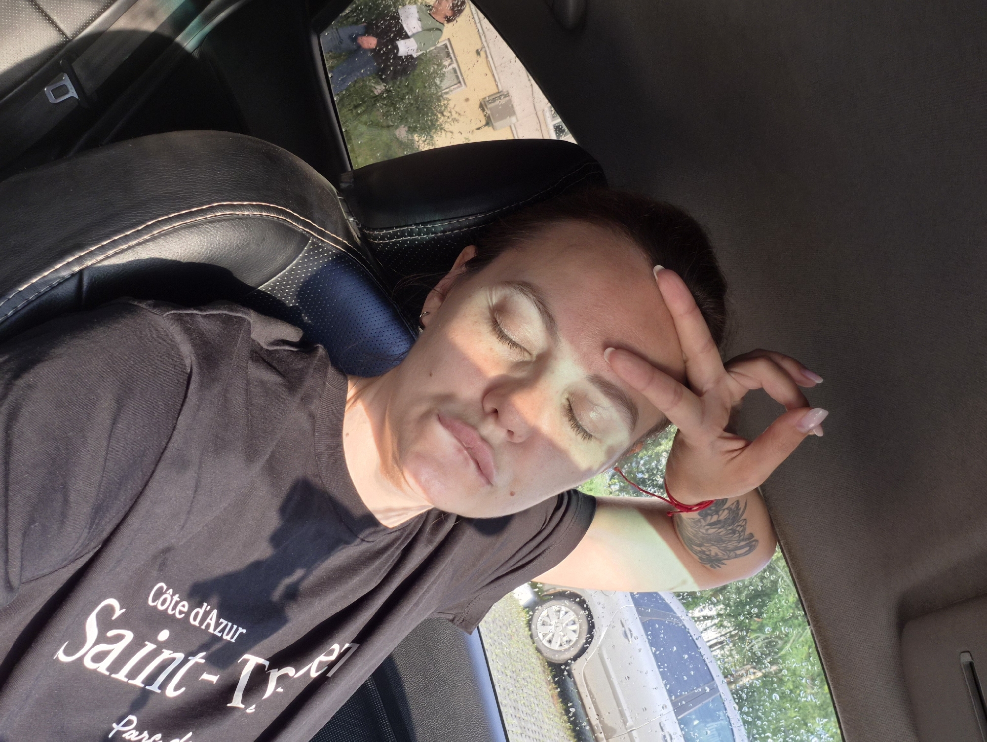 In Chaikovsky, Russia, a woman with a tattoo on her arm and wearing a black t-shirt with text is seen from the chest up, sitting in the passenger seat of a car. Her eyes are closed, and her hand is resting on her forehead. Sunlight is visible on her face. Through the car window, trees and a car are visible, as is a person standing outside. The interior of the car, including the black leather seat, is also visible.