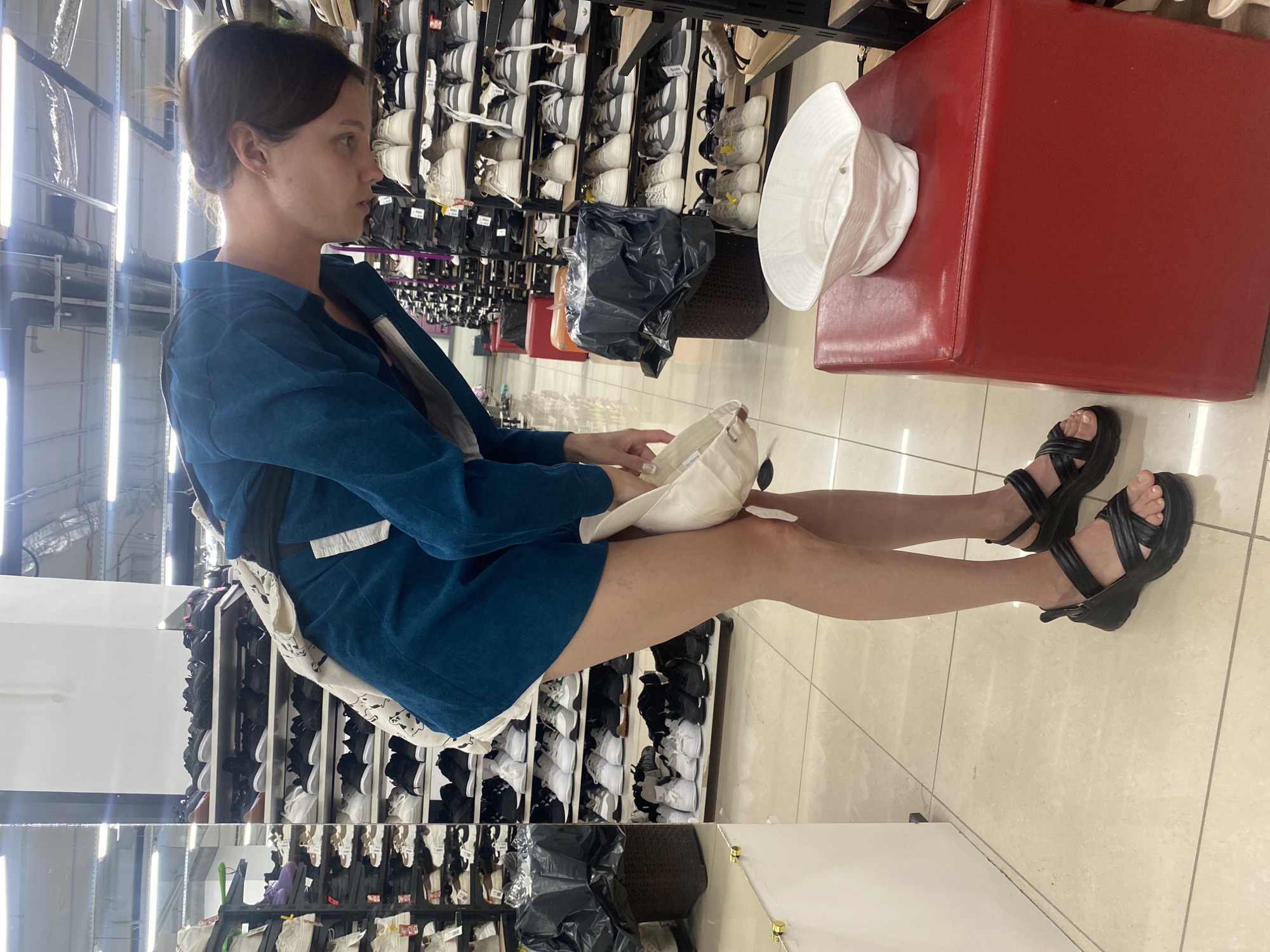 The image depicts a person in a shoe store in Novokuybyshevsk, Russia. The individual, wearing a teal jacket and black platform sandals, is bent over and appears to be examining a white bucket hat placed on a red ottoman. Rows of shoes are visible on shelves in the background, suggesting a retail environment. The floor is tiled, and fluorescent lighting illuminates the scene from above. A large black trash bag is situated next to a shelf of shoes on the left side of the image.