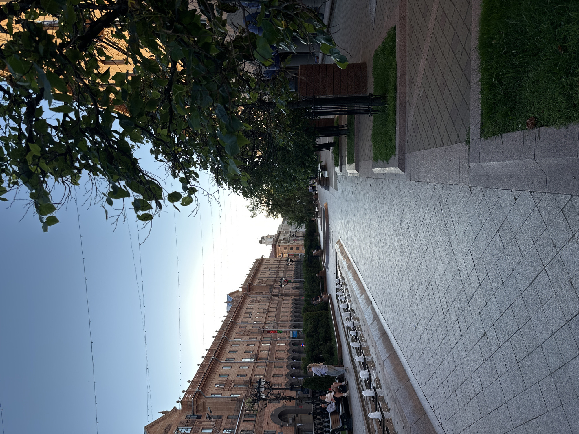 The image depicts a street scene in Minsk, Belarus, with a focus on a large, ornate building and a paved pedestrian walkway lined with a fountain and greenery. The building, constructed of reddish-brown brick, features numerous windows and arched entrances, suggesting an architectural style typical of the mid-20th century. Lights are visible along the building's facade and on lampposts lining the walkway.
On the walkway, a fountain with multiple spouts is active, with water spraying into the air. Several people are seated on benches near the fountain, and others are walking along the path. One person is standing near the fountain, seemingly observing it. The walkway is paved with light-colored tiles arranged in a diamond pattern. Adjacent to the walkway are strips of manicured green grass, bordered by stone.
In the foreground, a large tree with dark green leaves partially obscures the view, casting shadows on the scene. The sky is a clear, pale blue, indicating it is likely daytime, possibly late afternoon or early evening due to the soft light. The overall atmosphere appears calm and pleasant, with people enjoying the public space.
While no specific text is clearly legible on the building or signage, the architecture and the presence of a flag on the building (partially visible) are location cues. The scene suggests a leisurely day in an urban environment, with a blend of historical architecture and modern public amenities.