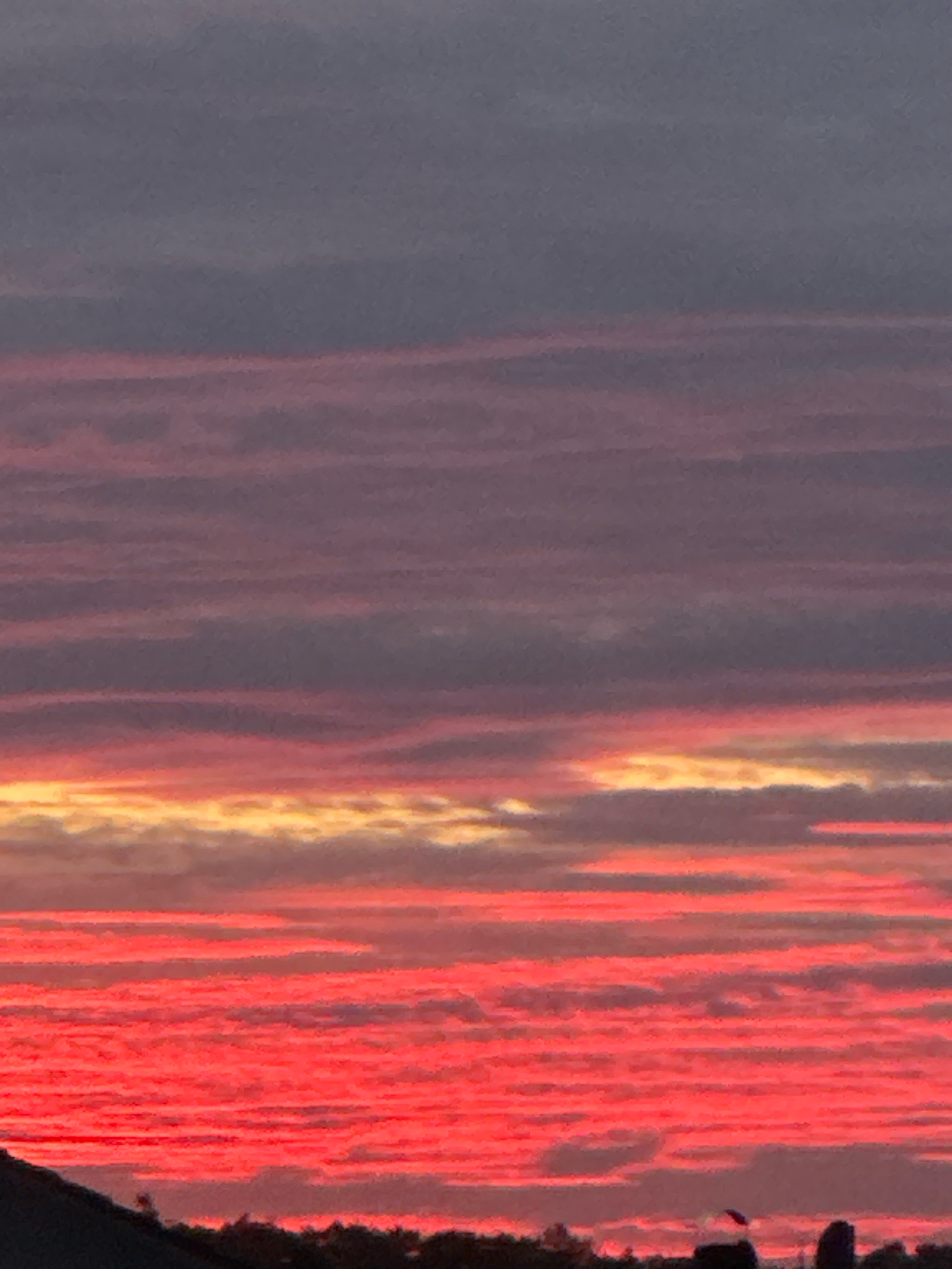 The image shows a sunset sky with horizontal bands of color. The sky is a gradient of colors, starting with deep gray at the top, transitioning to purple, then to pink and red hues. There are thin, darker horizontal lines within the colored bands, suggesting layers of clouds. The bottom of the image shows a silhouette of trees and some indistinct structures against the bright sky, possibly indicating a landscape or cityscape. The scene appears to be an outdoor environment at dusk. Based on the provided context, this sunset was photographed in Wettstetten, Germany.