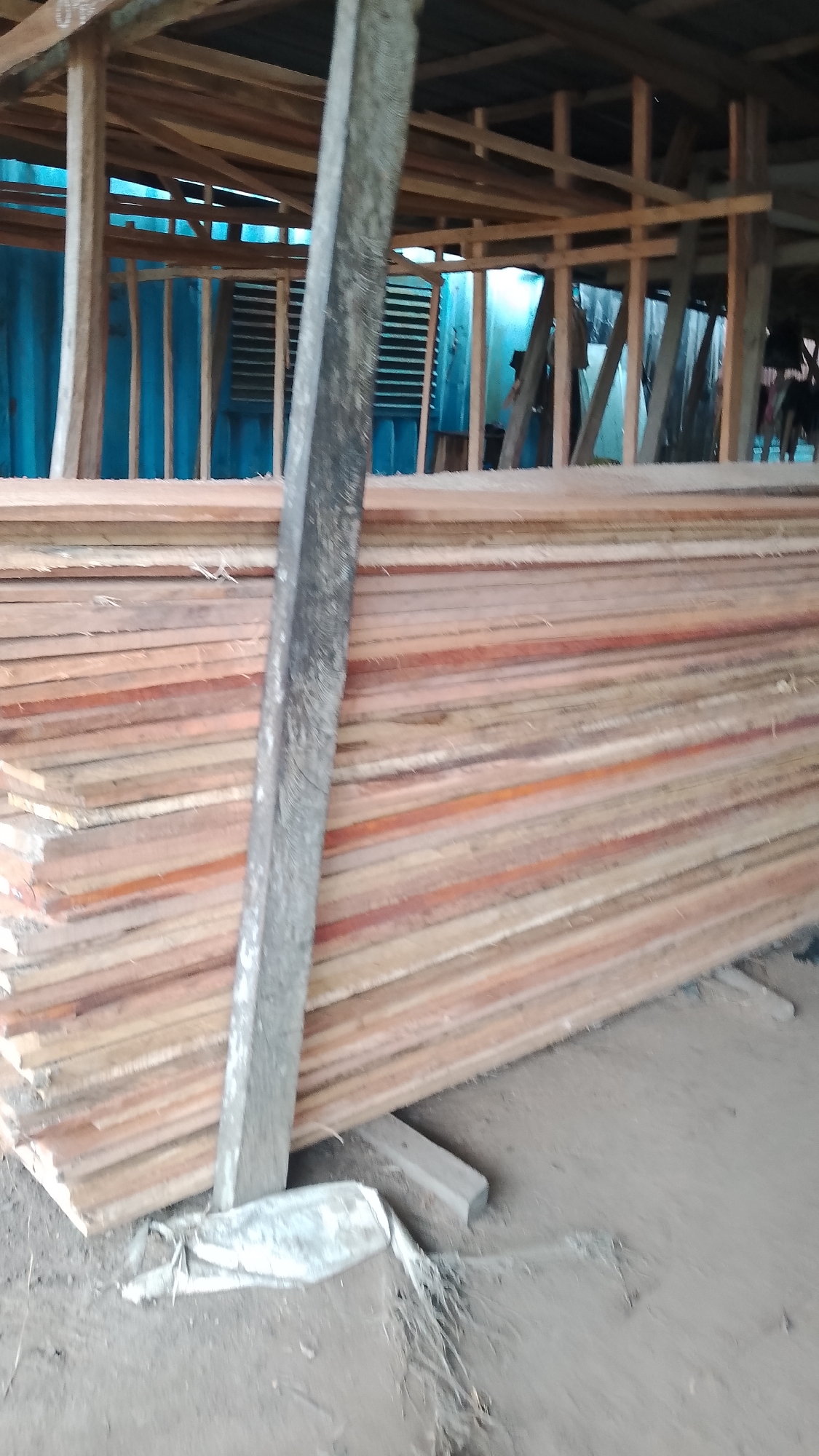 A stack of lumber rests indoors in Asubi, Nigeria. The lumber appears to be stacked on the ground with pieces of wood supporting it. A weathered, square wooden post leans against the stack. The structure around the lumber is wooden, with a corrugated metal roof. A person can be seen behind the lumber near a blue corrugated metal wall. The ground is dirt.