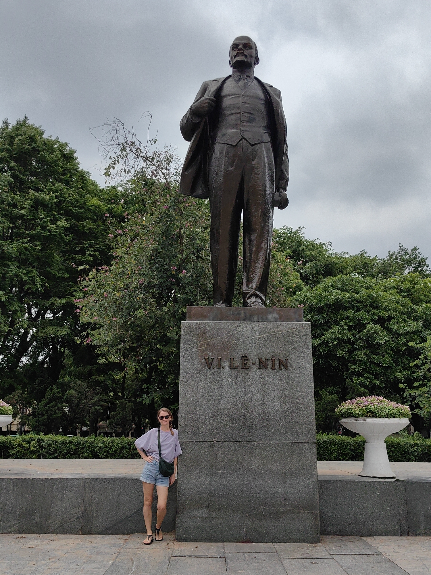 The image captures a large bronze statue of V.I. Lê-nin (Vladimir Lenin) standing on a tall, gray stone pedestal in an outdoor public space in Hà Nội, Vietnam. The statue portrays Lenin in a suit and waistcoat, with his left hand holding the lapel of his jacket. The inscription "V.I. LÊ-NIN" is prominently displayed in raised bronze letters on the front of the pedestal.
To the left of the imposing monument, a woman is posing for the camera. She stands casually, leaning against the pedestal's base, with her left hand on her hip. She wears a light purple short-sleeved top, denim shorts, black flip-flops, and sunglasses, carrying a dark green cross-body bag. Her expression is relaxed, featuring a slight smile.
The background is filled with lush green foliage, including tall trees and flowering bushes with small pink blossoms, indicative of a tropical or subtropical climate. The sky above is overcast with gray clouds, suggesting a cloudy daytime. On the right side of the statue's base, a white, goblet-shaped concrete planter holds a vibrant arrangement of small pink and purple flowers. The ground is paved with large, light gray flagstones. The woman's presence provides a sense of scale, highlighting the considerable height and grandeur of the statue.