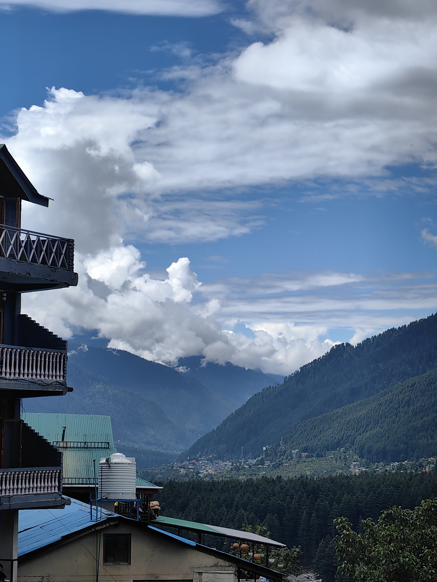 The image depicts a mountainous landscape in the city of Vashisht, India, under a partly cloudy sky. In the foreground, a multi-story building with balconies is visible on the left side. The building has a dark exterior and white balustrades. Below this building, parts of other structures with blue and green roofs are also present, along with a large white water tank.
The midground features a valley with a town nestled on the slopes of a mountain, characterized by numerous buildings with red and multi-colored roofs. This is surrounded by dense green forests. The background is dominated by rolling mountains, with the furthest peaks shrouded in a hazy, muted blue. The sky above is a vibrant blue, interspersed with large, puffy white clouds and thinner, streaky clouds. The overall scene conveys a sense of a hillside settlement within a vast natural environment.