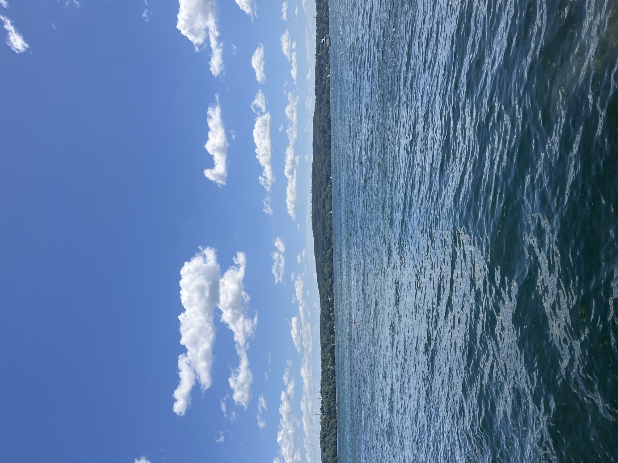 The image shows a wide view of Starnberger See, Germany. The upper portion of the image is dominated by a bright blue sky with scattered white cumulus clouds. Below the sky is a green, tree-covered hillside that forms the shoreline of the lake. The water in the foreground is a deep blue, with small waves and ripples indicating a gentle breeze. In the distance, along the shoreline, are scattered buildings and trees, suggesting a populated area. There are several sailboats visible on the water, and a larger white boat is cruising in the distance. A single orange buoy is also present in the water. The overall scene is one of a calm, sunny day on a large lake.