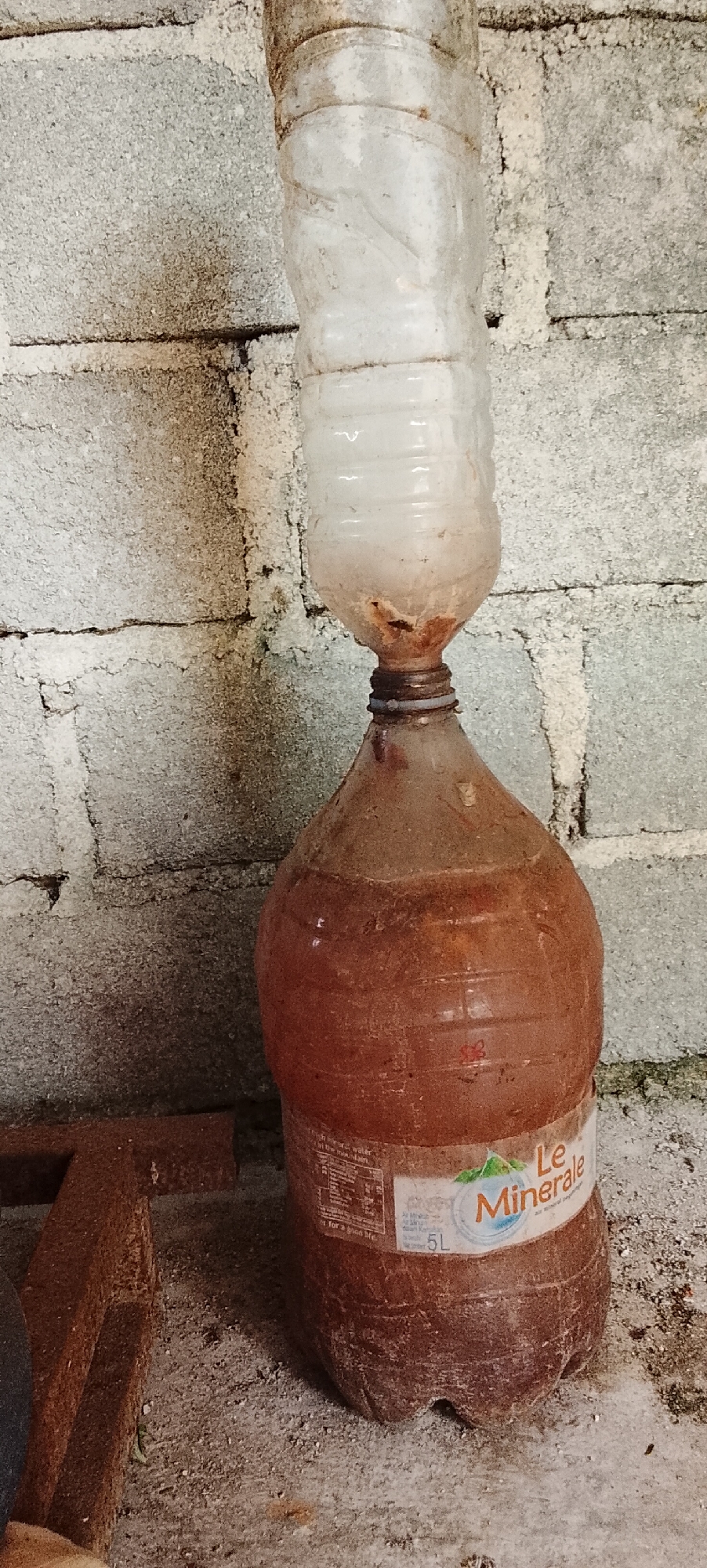 The image shows two stacked plastic bottles against a grey cinder block wall. The top bottle, which appears to be a cut-off section of a larger bottle, is inverted and placed inside the neck of the bottom bottle. The top portion of the bottle is translucent and shows some dried residue. The bottom bottle is larger and has a reddish-brown liquid inside. A label on the bottom bottle reads "Le Minerale" and indicates "5L". The bottles are dusty, and the surrounding environment appears to be a somewhat unkempt or utilitarian space, possibly outdoors or in a storage area, given the cinder block construction and what appears to be a piece of rusty metal object in the lower left corner. The image does not contain any information that would suggest its location in the city of Padang Dalom, Indonesia.