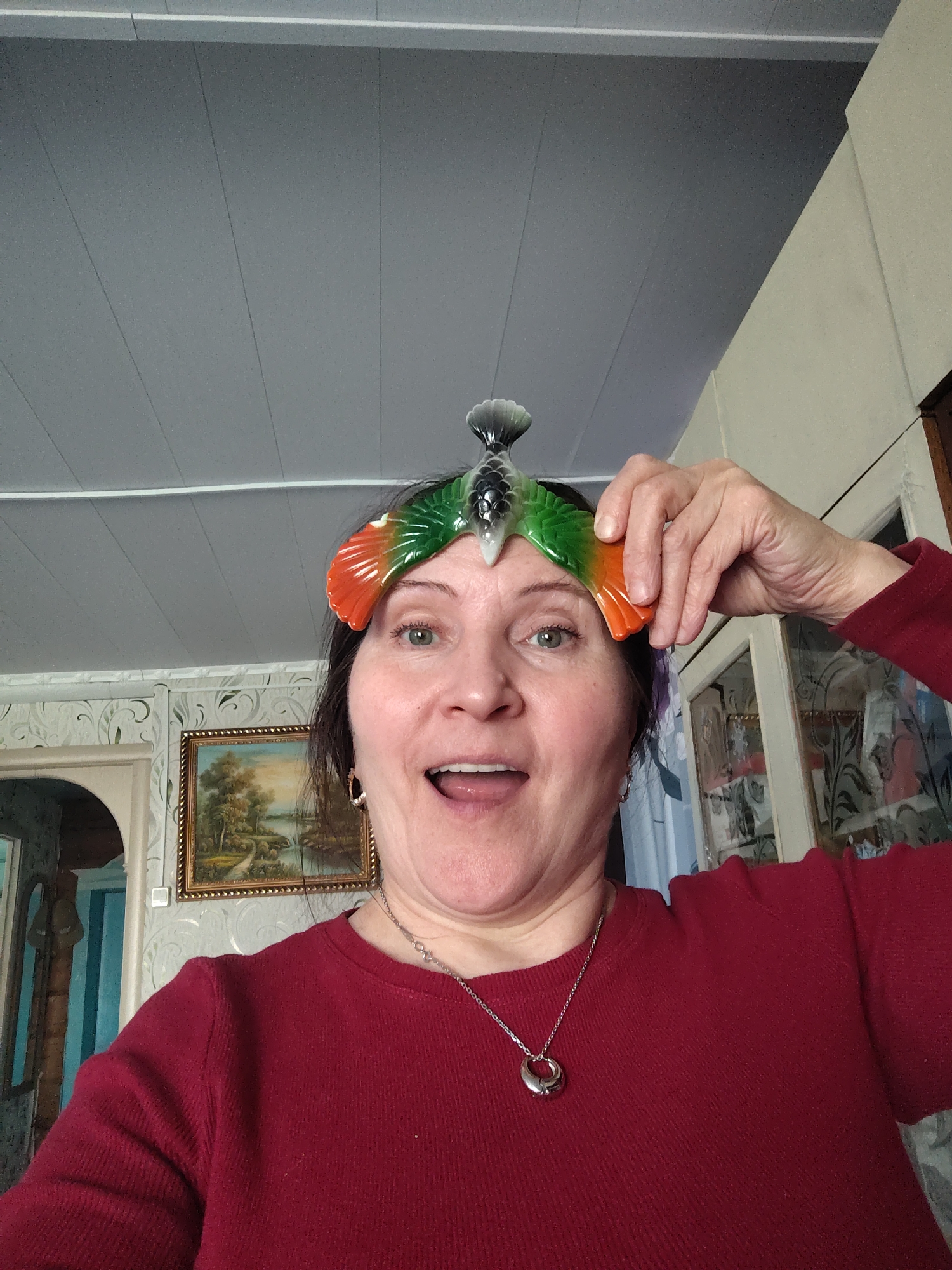 The image presents a candid, close-up portrait of a middle-aged woman captured in a selfie-style indoors, likely in her home in Сусады-Эбалак, Russia. She looks directly at the camera with a wide-eyed, open-mouthed expression of amusement, surprise, or joy. Her dark hair frames her face, and she wears a simple red long-sleeved top and a silver chain necklace with a distinctive circular pendant.
A prominent detail is the colorful, decorative object perched on her head, which she supports with her right hand. This item appears to be a bird or fish figurine, featuring translucent green and orange sections resembling wings or fins, and a grey, textured tail.
The background elements suggest a domestic setting. Above the woman, a light grey ceiling with visible panels and white strips is visible. Behind her, a wall is covered with light-colored wallpaper featuring a subtle white scroll pattern. A gold-framed landscape painting, depicting a calm scene with trees and a body of water, hangs on this wall. To her right, part of a wooden cabinet with glass doors is visible. To her left, an arched doorway or opening, adorned with blue and white striped wallpaper, can be seen. The lighting suggests it is daytime. There is no visible text in the image. The woman's playful expression is the most notable emotional cue.